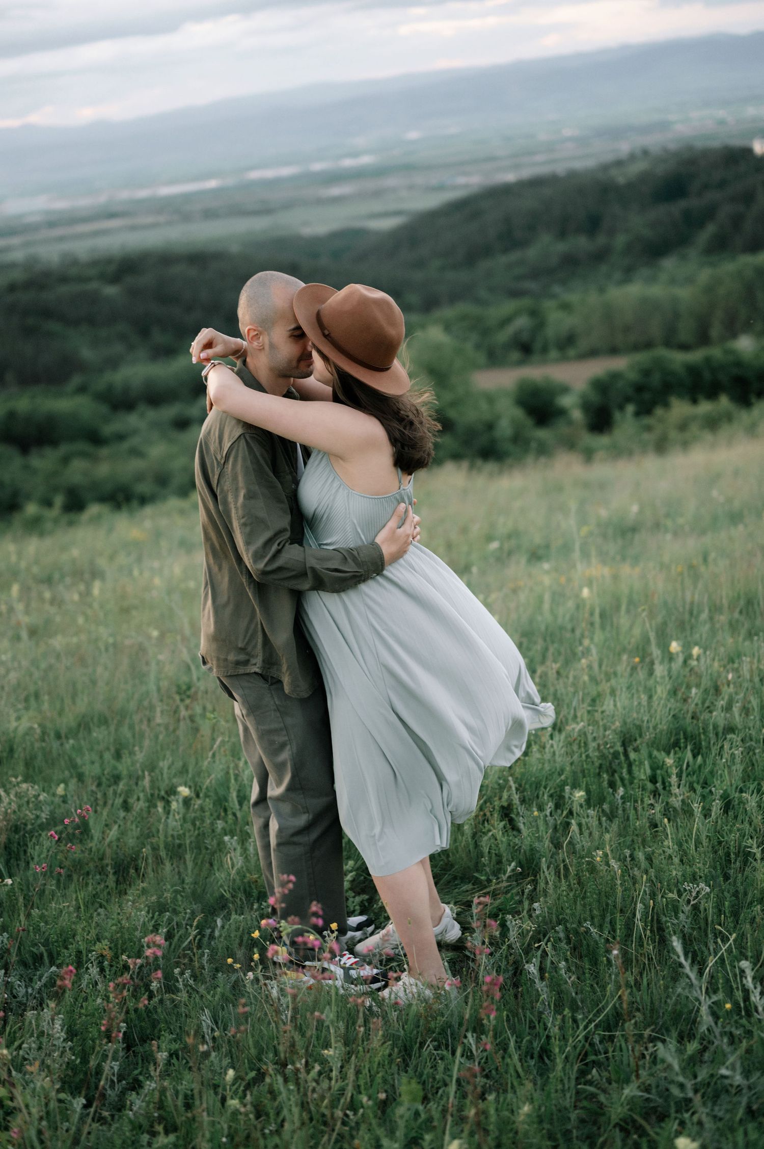 A couple embraces romantically in a green meadow, woman in light dress and brown hat, scenic valley in background.