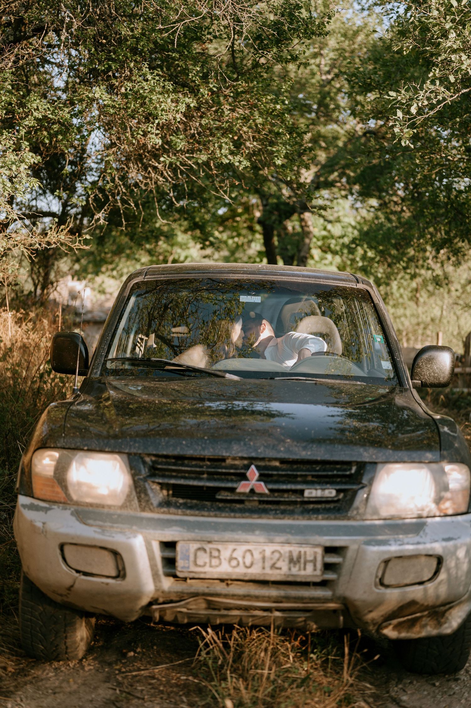 Abandoned Mitsubishi vehicle with headlights on parked under trees in a rural setting.