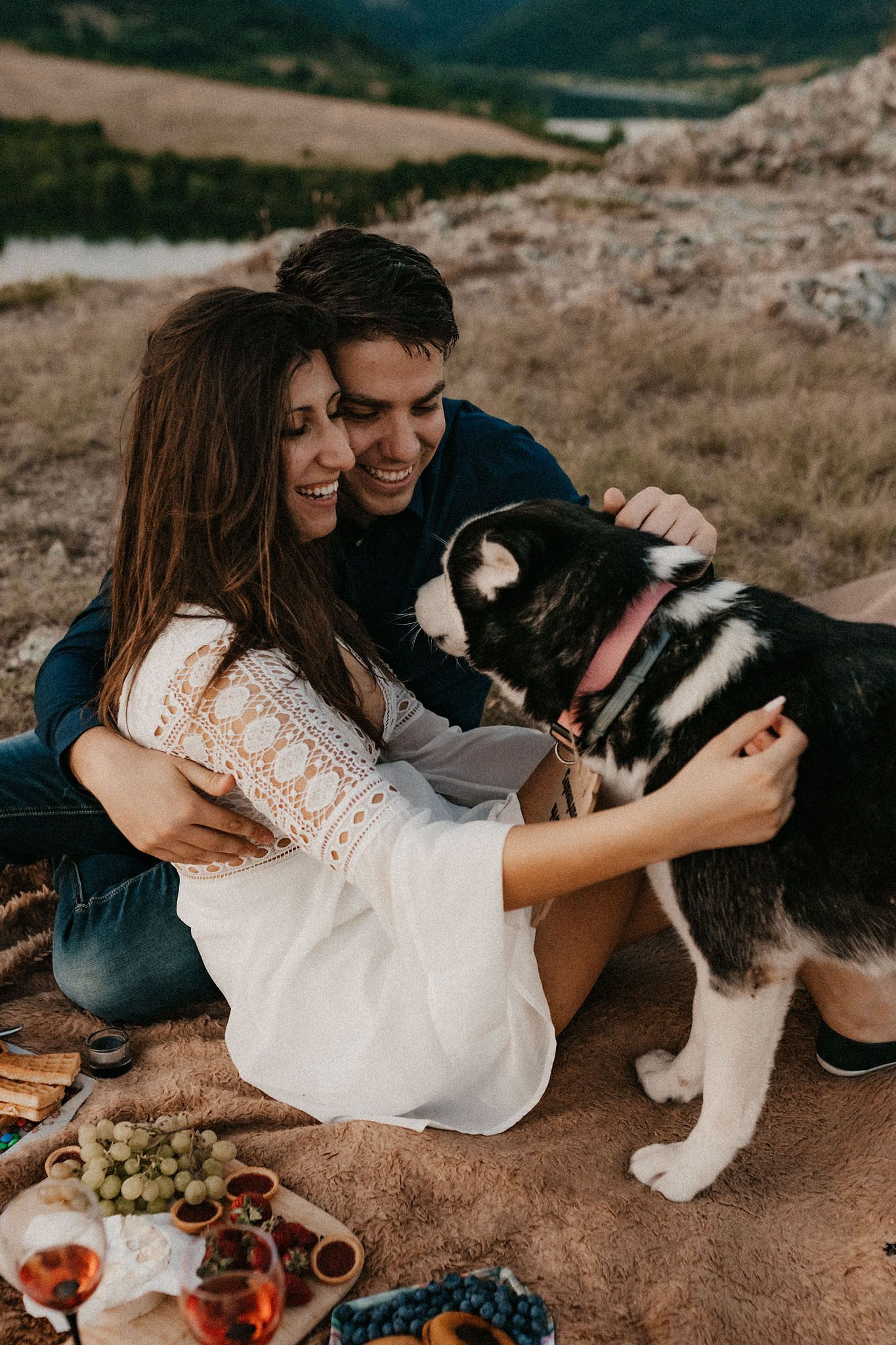 Couple cuddling with their husky dog during a scenic mountaintop picnic.