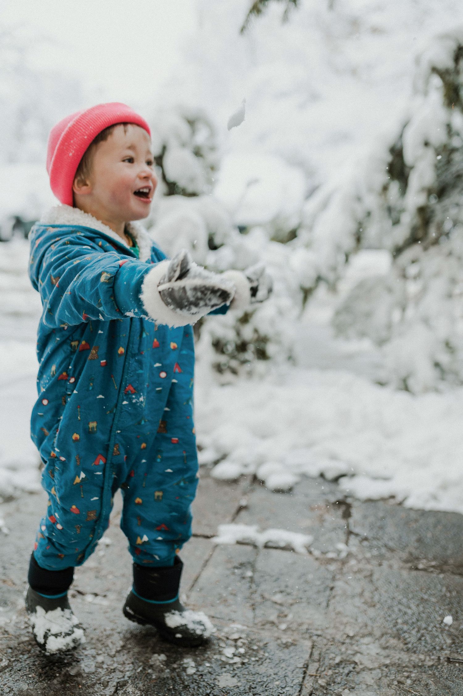 A small child in a teal snowsuit and pink hat joyfully plays in the winter snow.