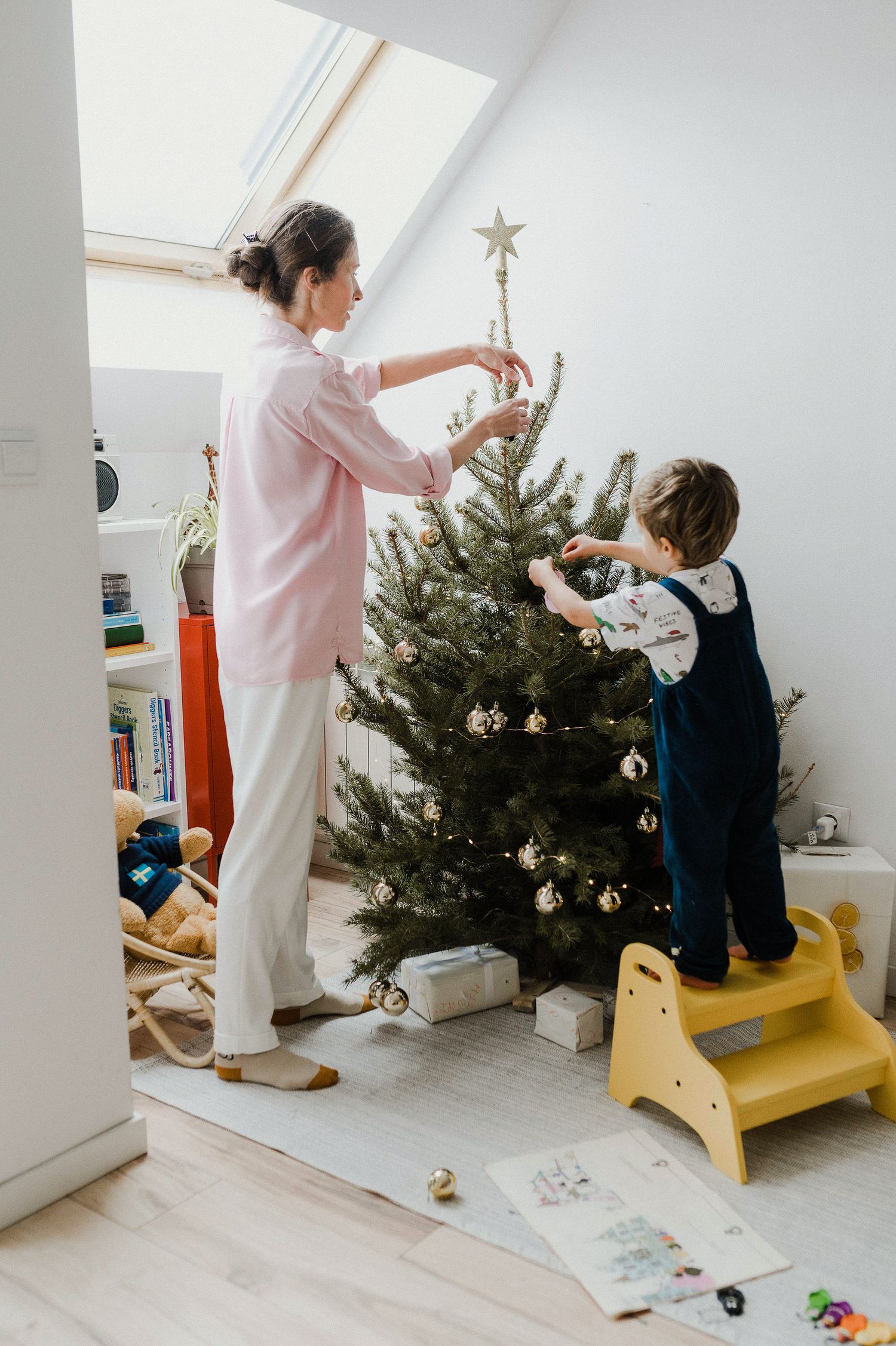Two people decorate a Christmas tree together in a bright attic room with sloped ceilings and skylights.