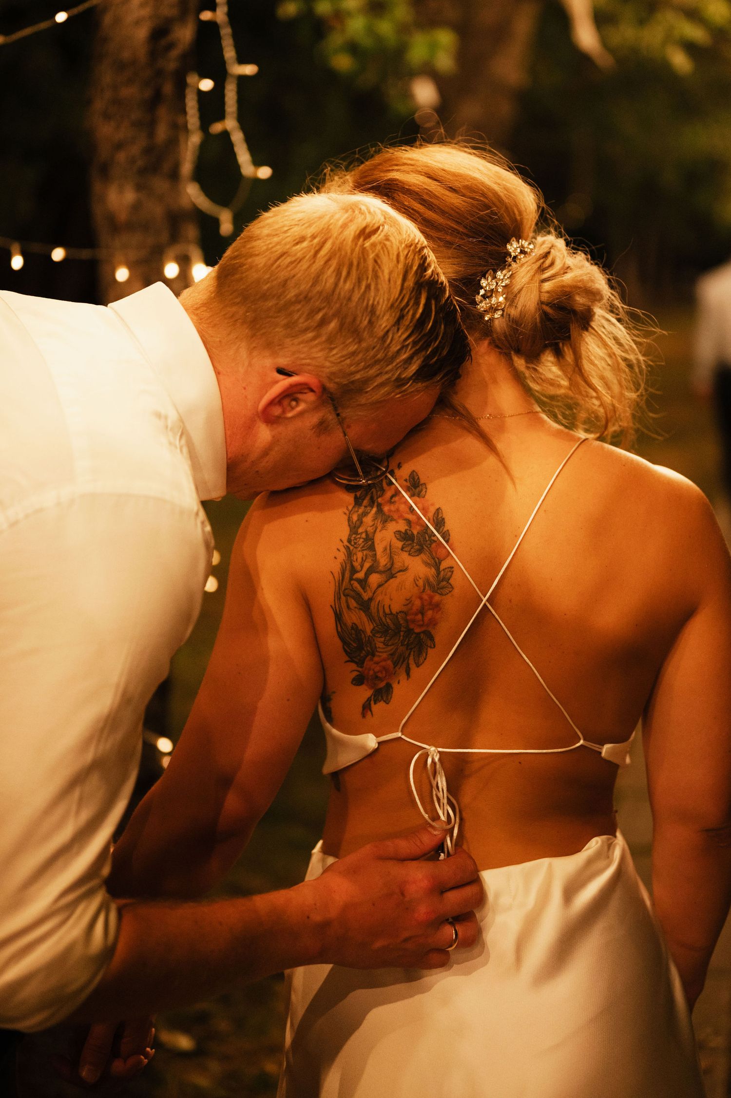 An intimate wedding moment captured outdoors with string lights and a detailed back tattoo visible.