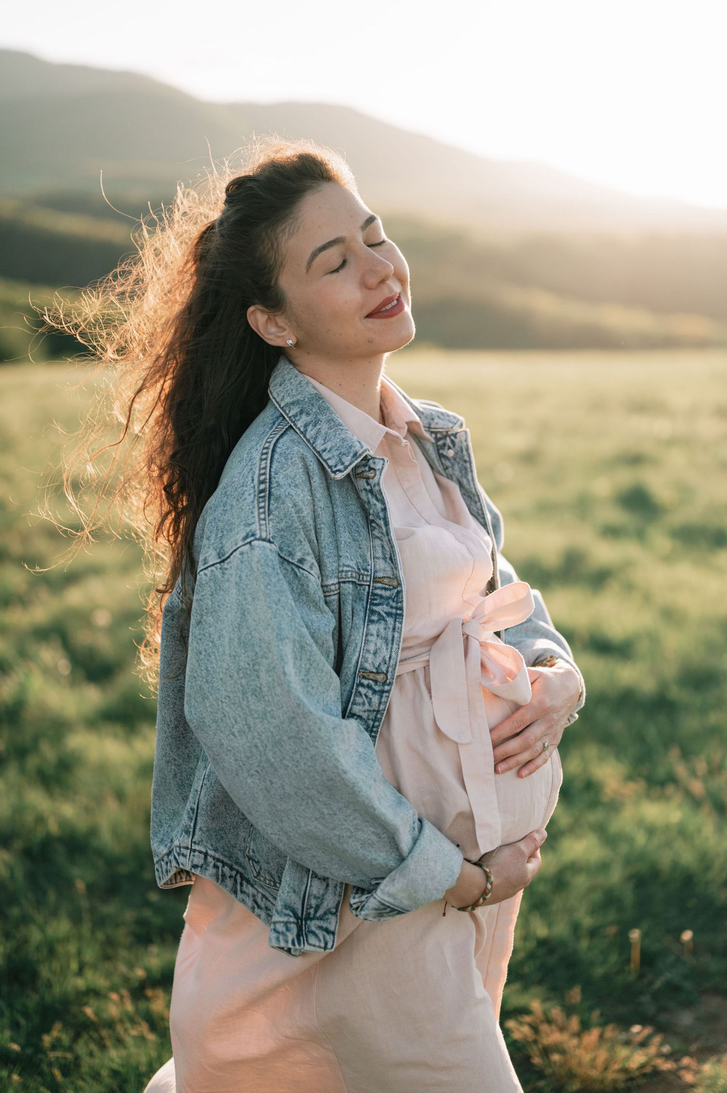 Woman in denim jacket and pink dress poses in sunlit mountain meadow.
