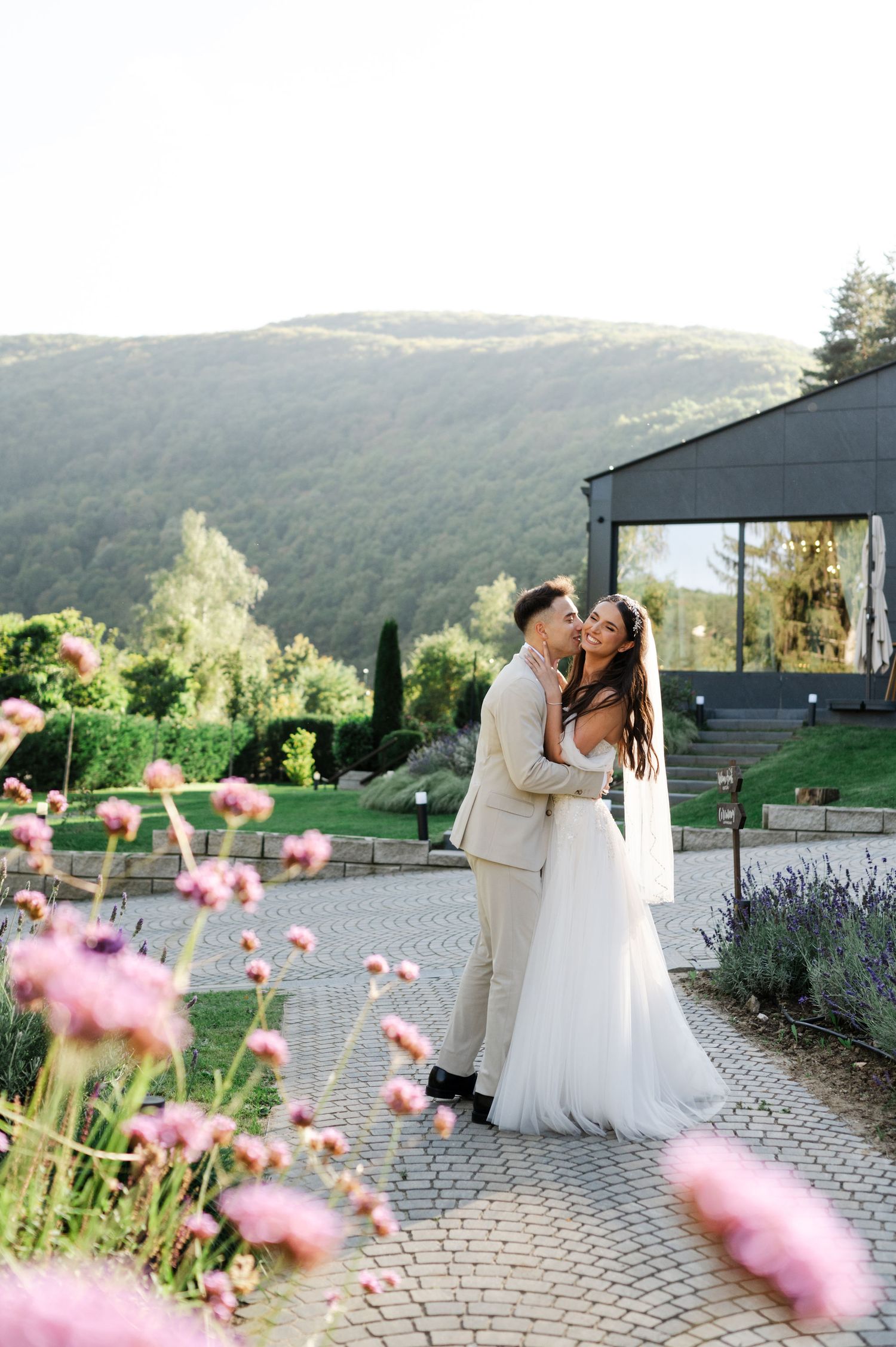 A romantic wedding couple shares a tender moment on a stone patio with pink flowers and mountain views at sunset.