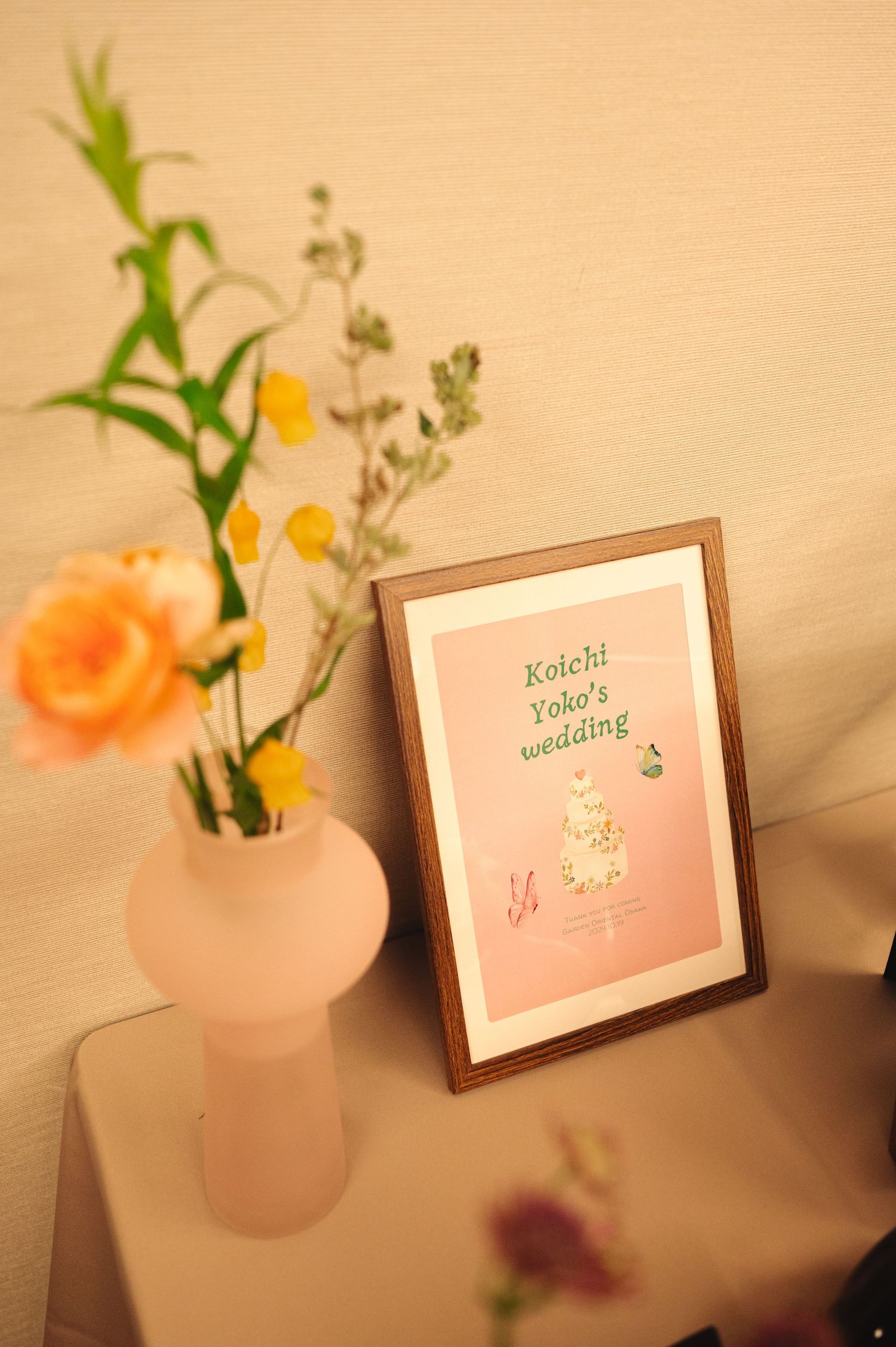 Pink vases with fresh flowers next to framed wedding signage on a light-colored surface.