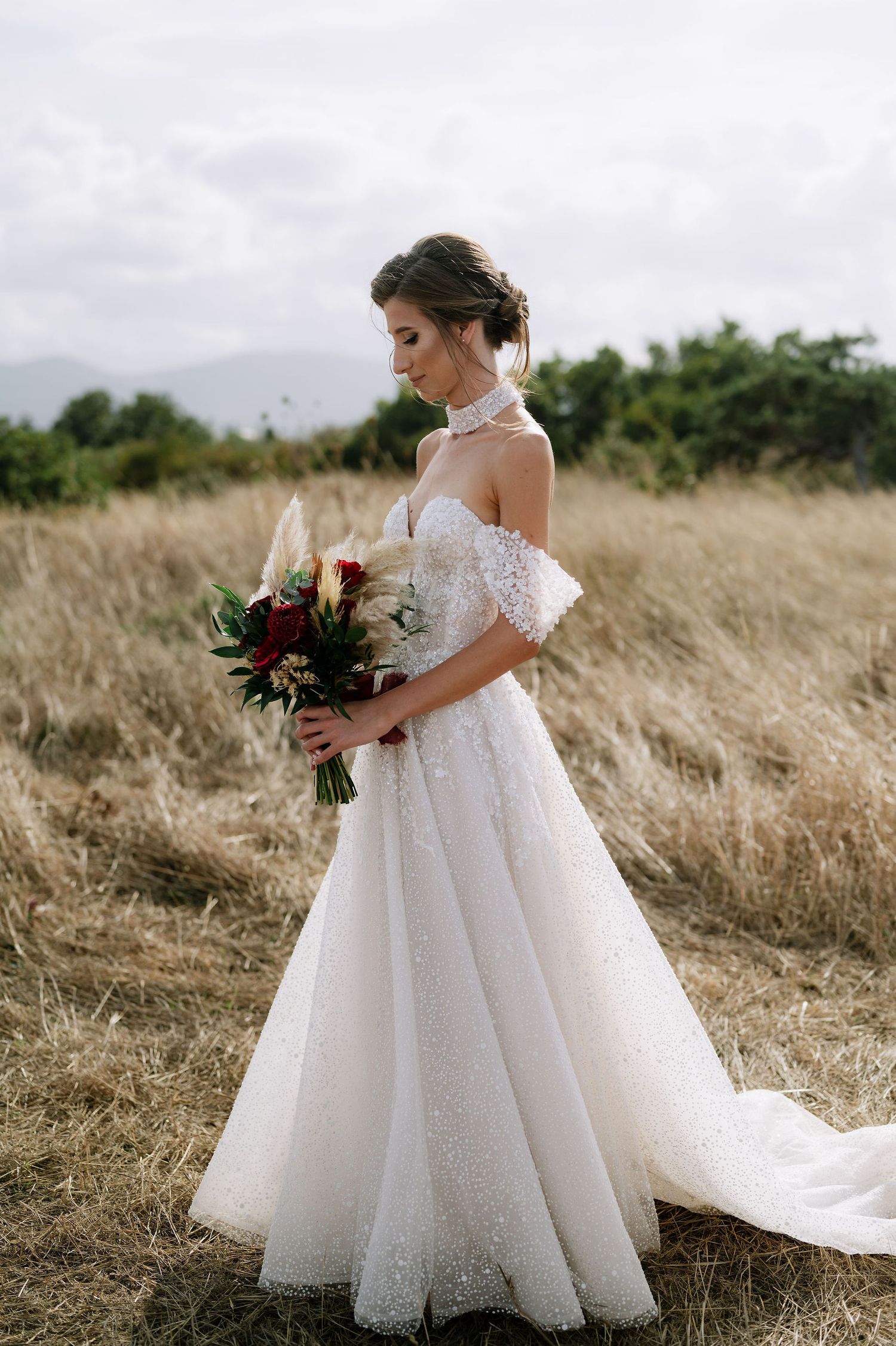 A bride in a flowing white ballgown wedding dress stands in a golden field holding a burgundy bouquet.