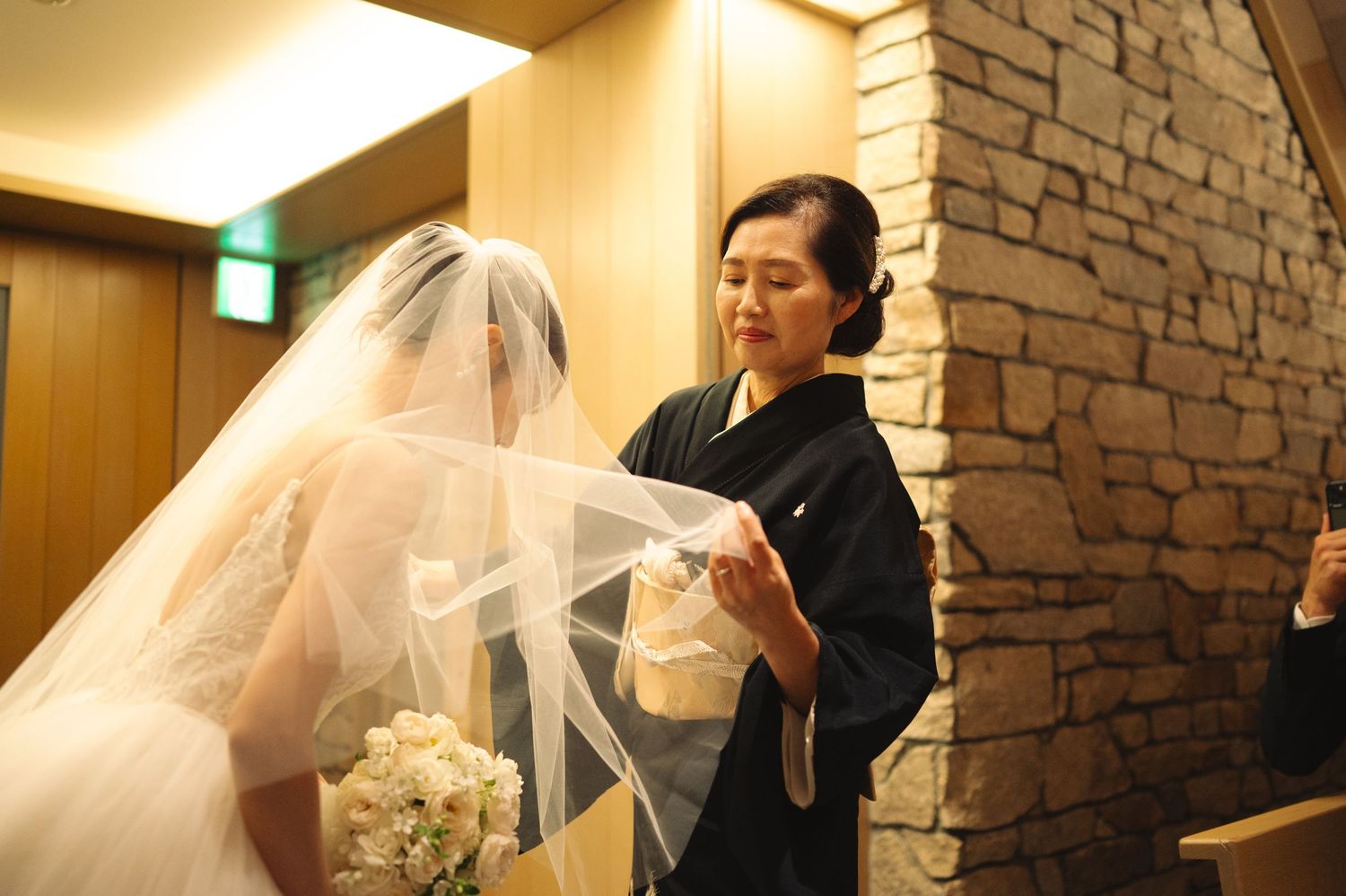 Bride in white gown receives final touches on her wedding dress from an attendant in black.