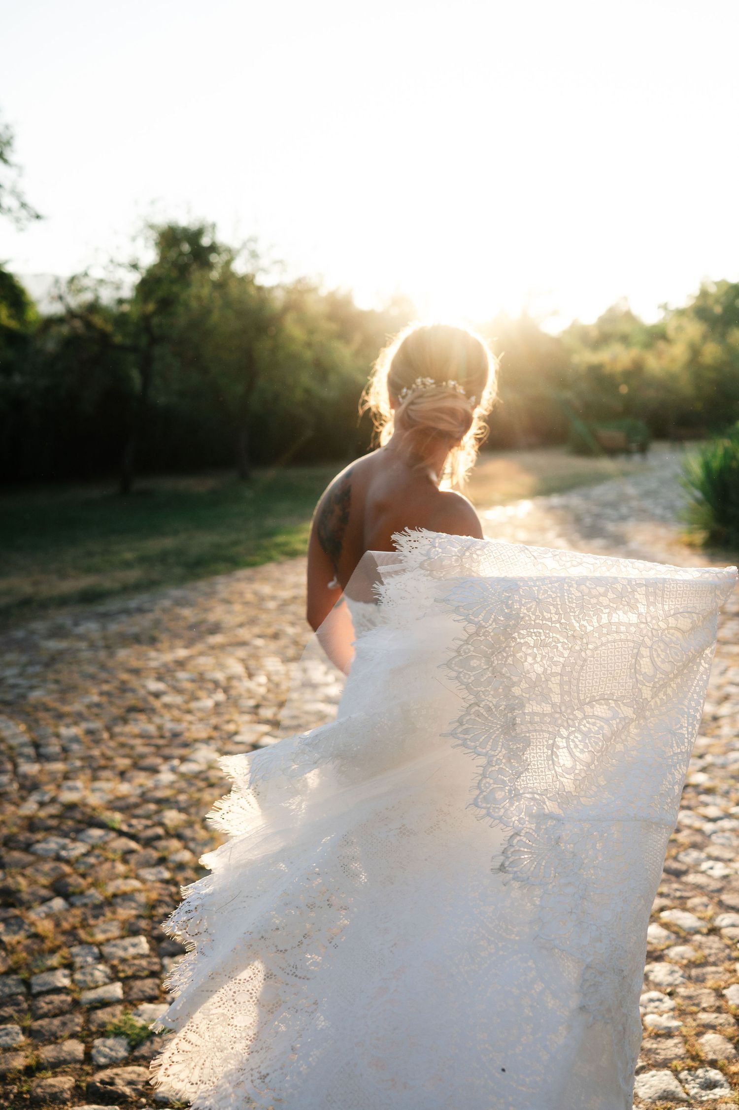 Ethereal bridal portrait with flowing lace dress catching the warm evening light on stone ground.