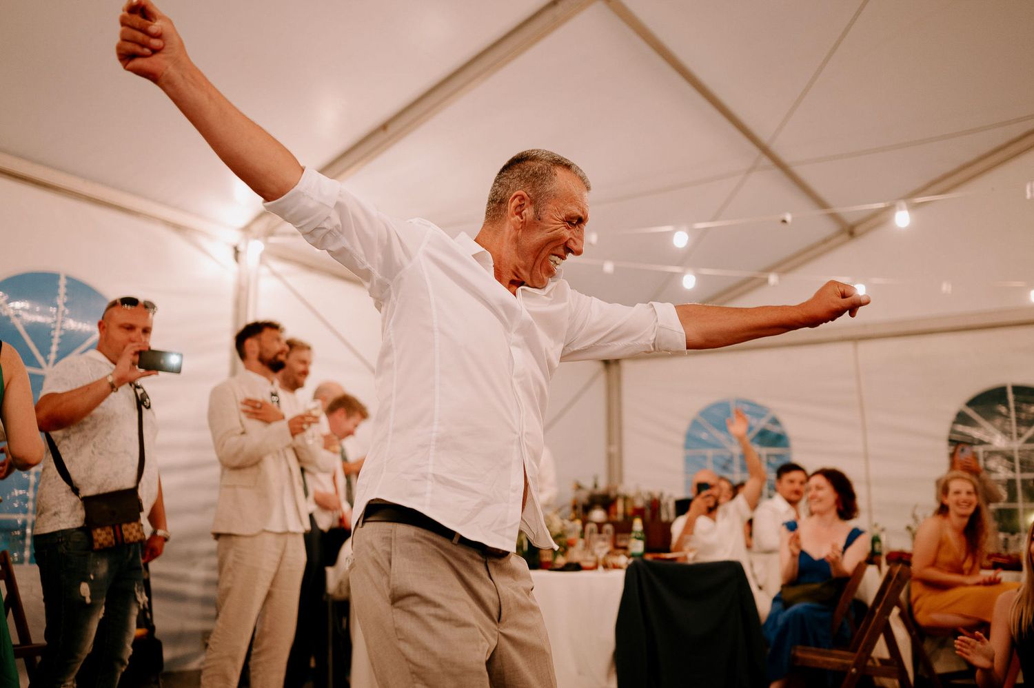 Guests celebrate and dance during a lively wedding reception under a white tent.