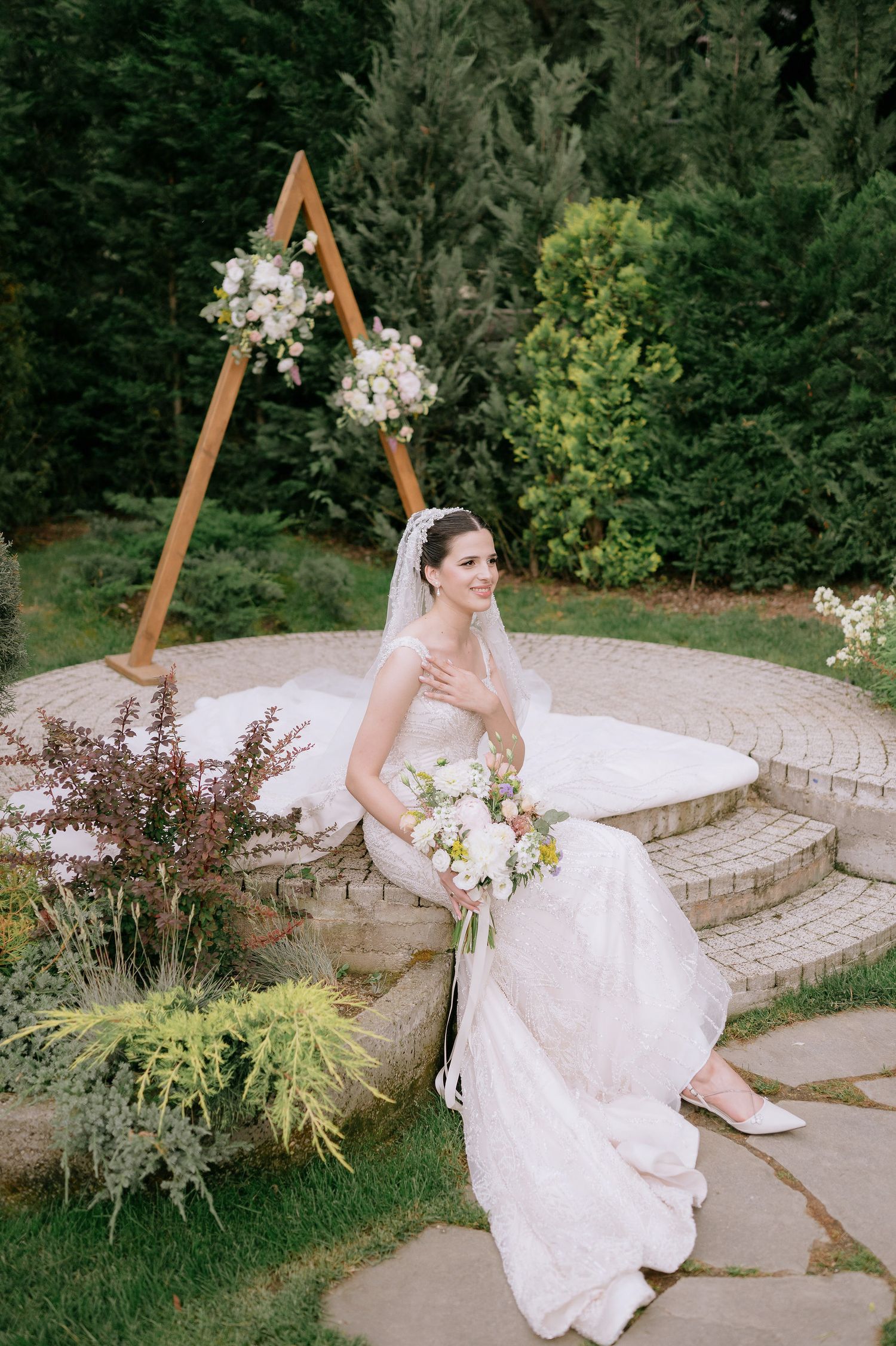 A bride in a flowing white dress sits elegantly on curved stone steps in a garden setting.