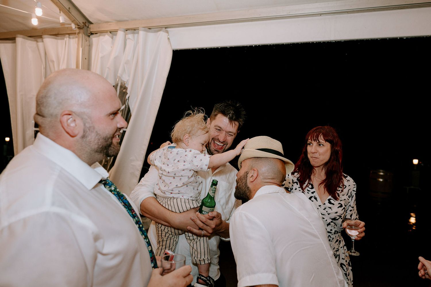 Wedding celebration with guests dancing and mingling in an illuminated tent space.