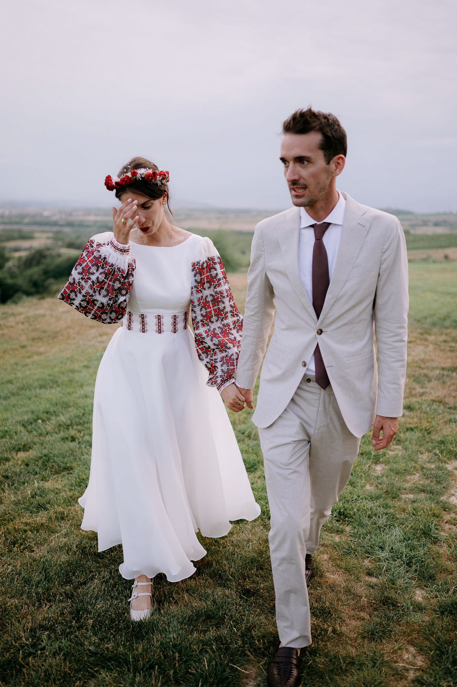 Wedding couple walks hand in hand through meadow wearing folk-inspired dress and light suit.
