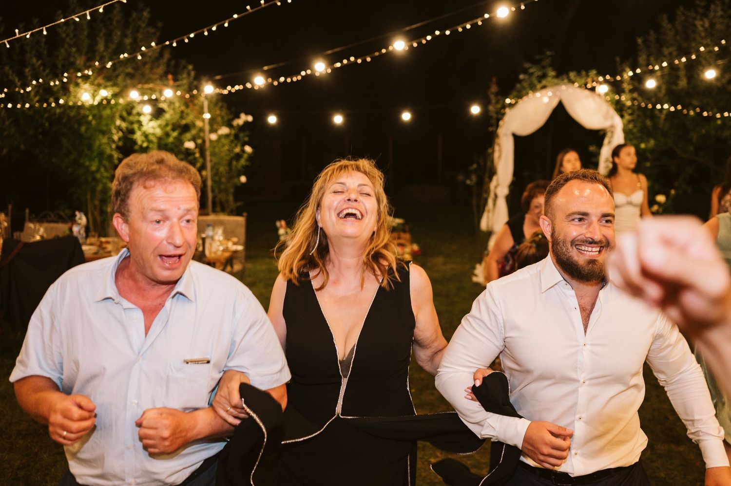 Wedding guests laughing and dancing at an outdoor reception under string lights.