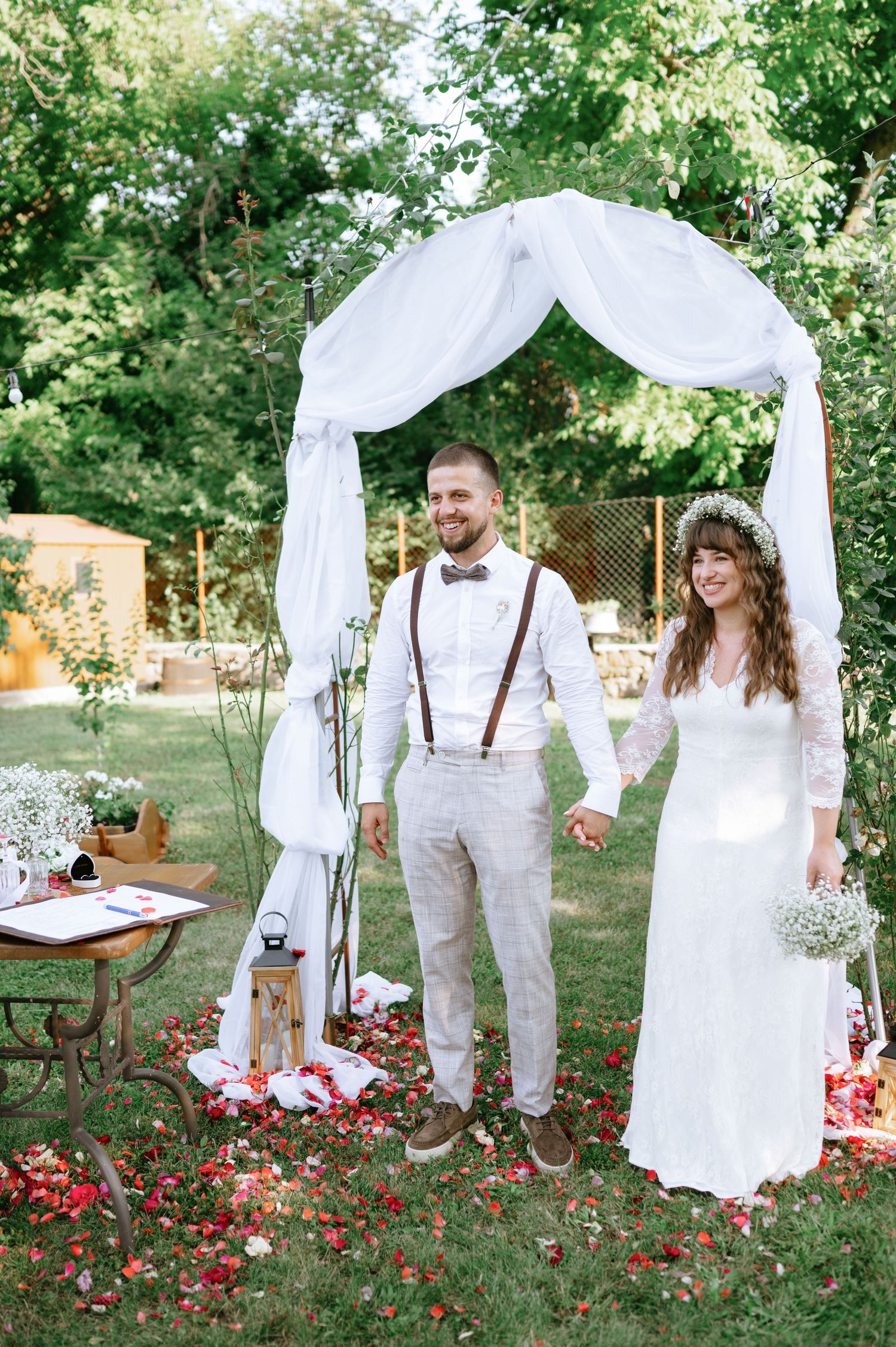 Romantic outdoor garden wedding setup with white arch, vintage furniture, and red flower petals scattered on grass.