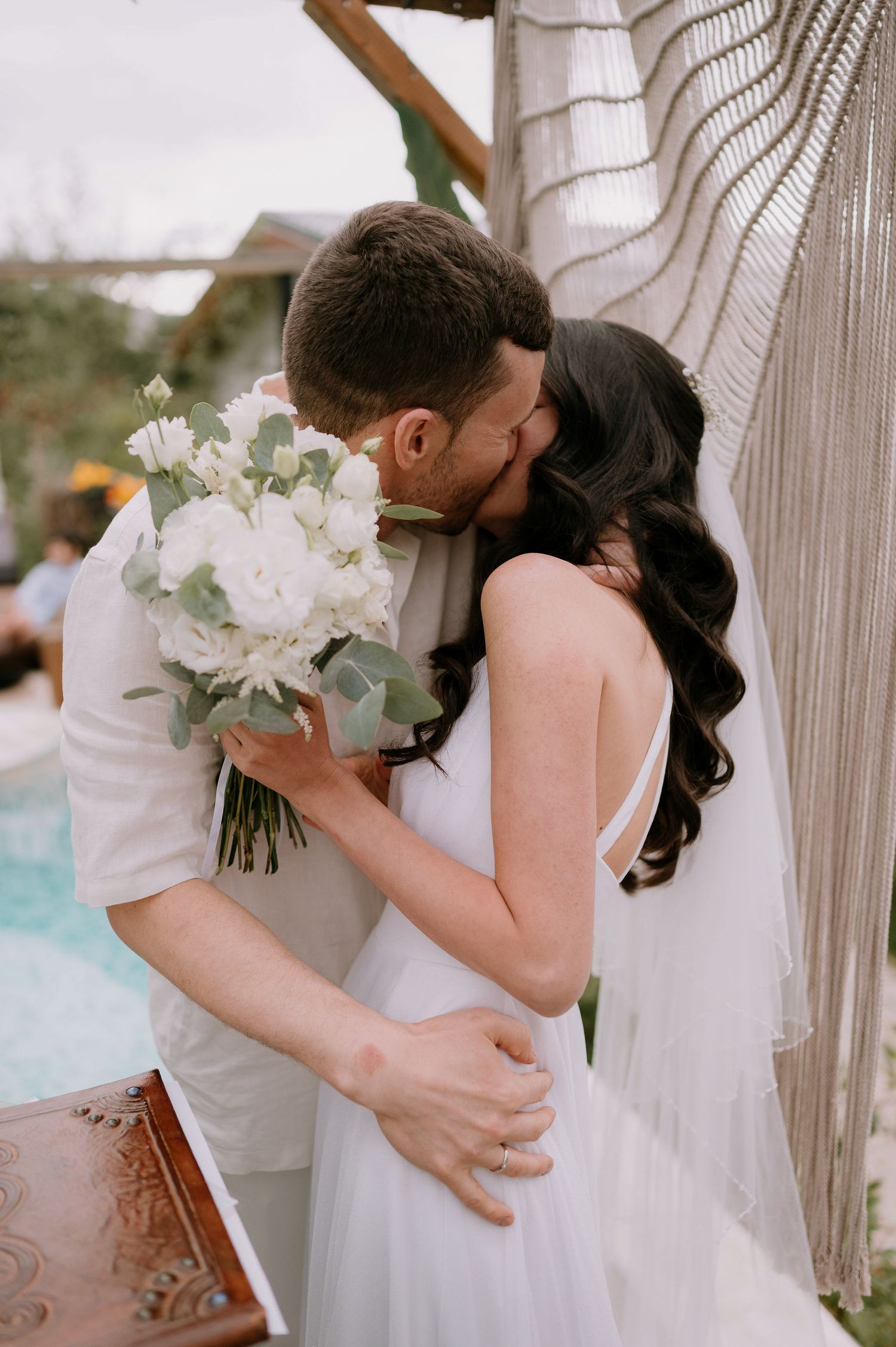 Intimate wedding moment with white bouquet and macrame backdrop by pool.