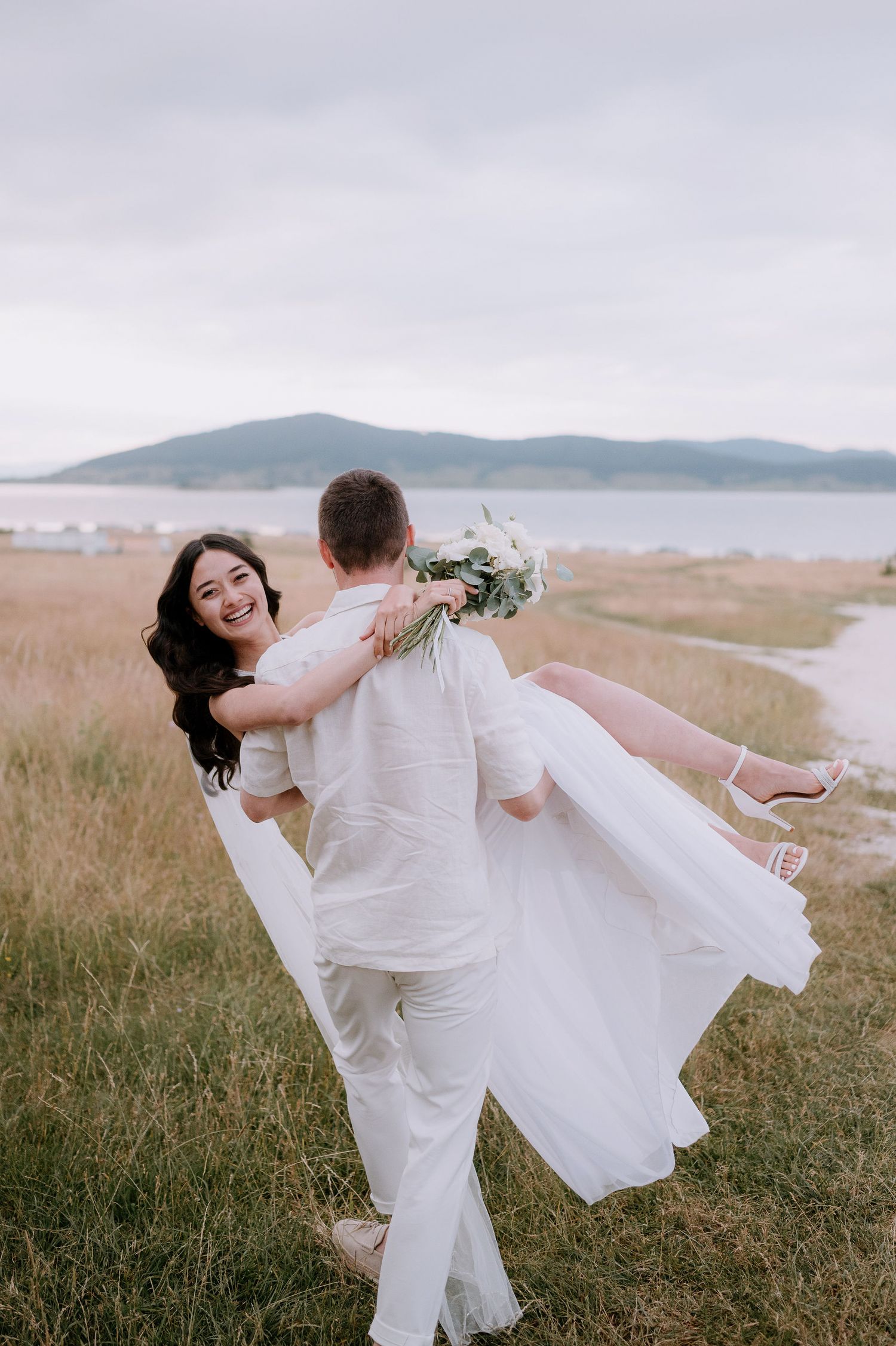 Newlyweds share a joyful dance moment in a scenic coastal field with mountains visible in the distance.