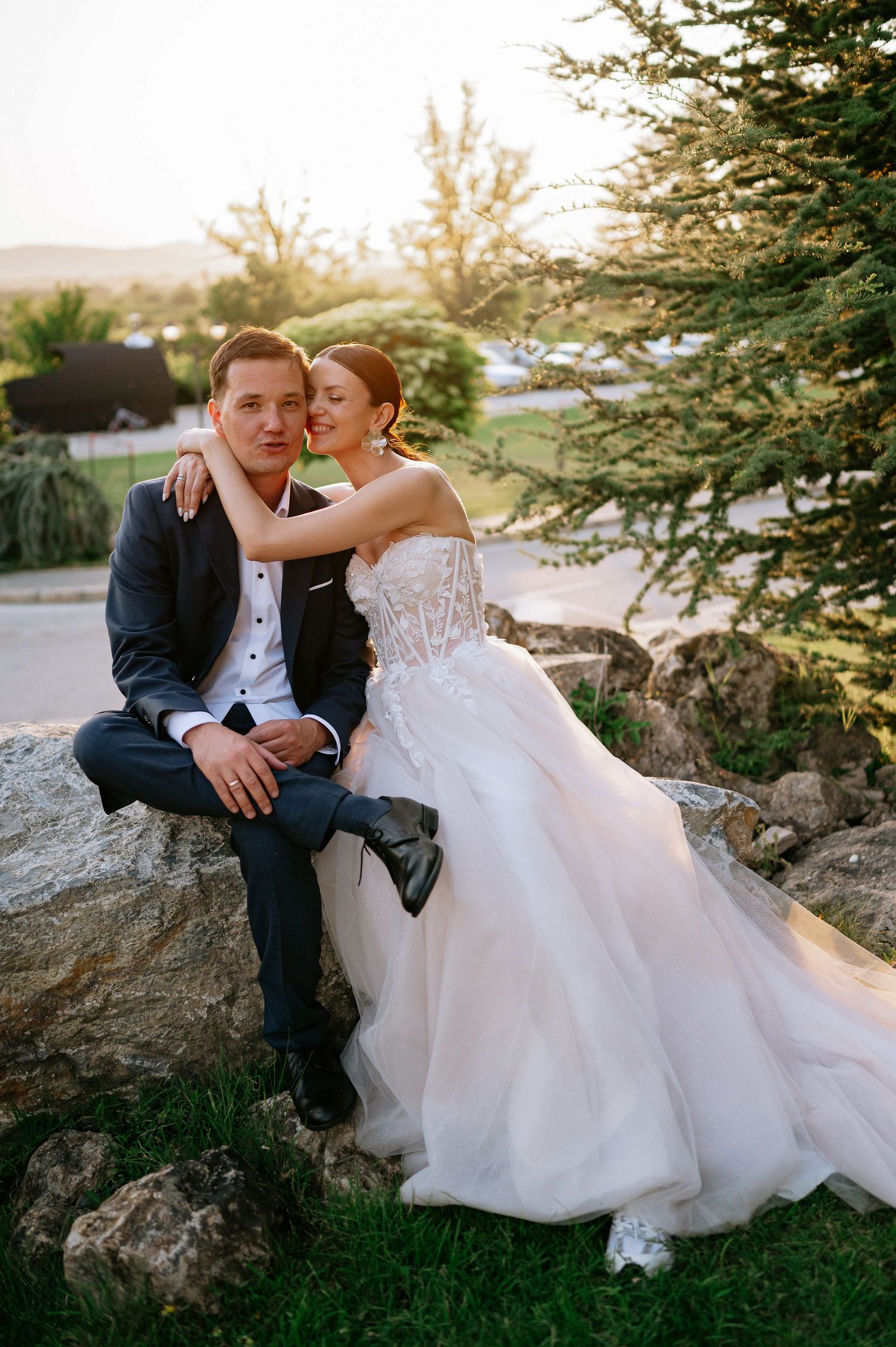 Joyful wedding couple shares laughs while sitting on stone landscaping at golden hour.