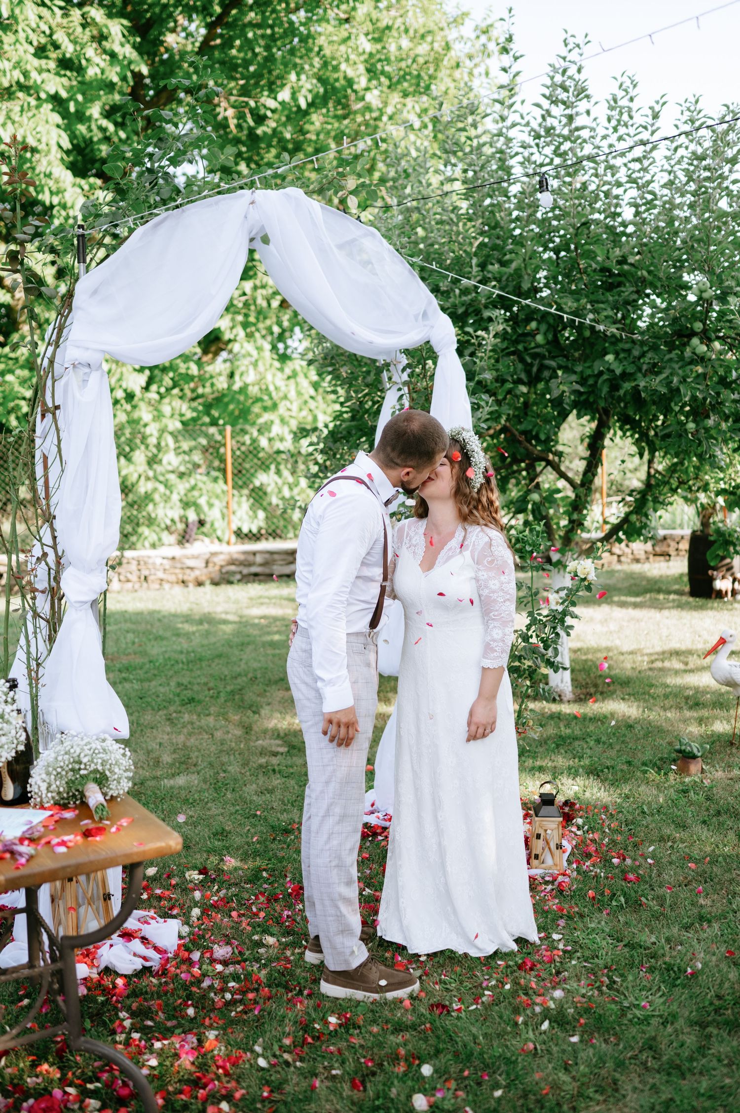 A romantic outdoor wedding ceremony with white draped arch and red flowers scattered on the grass.