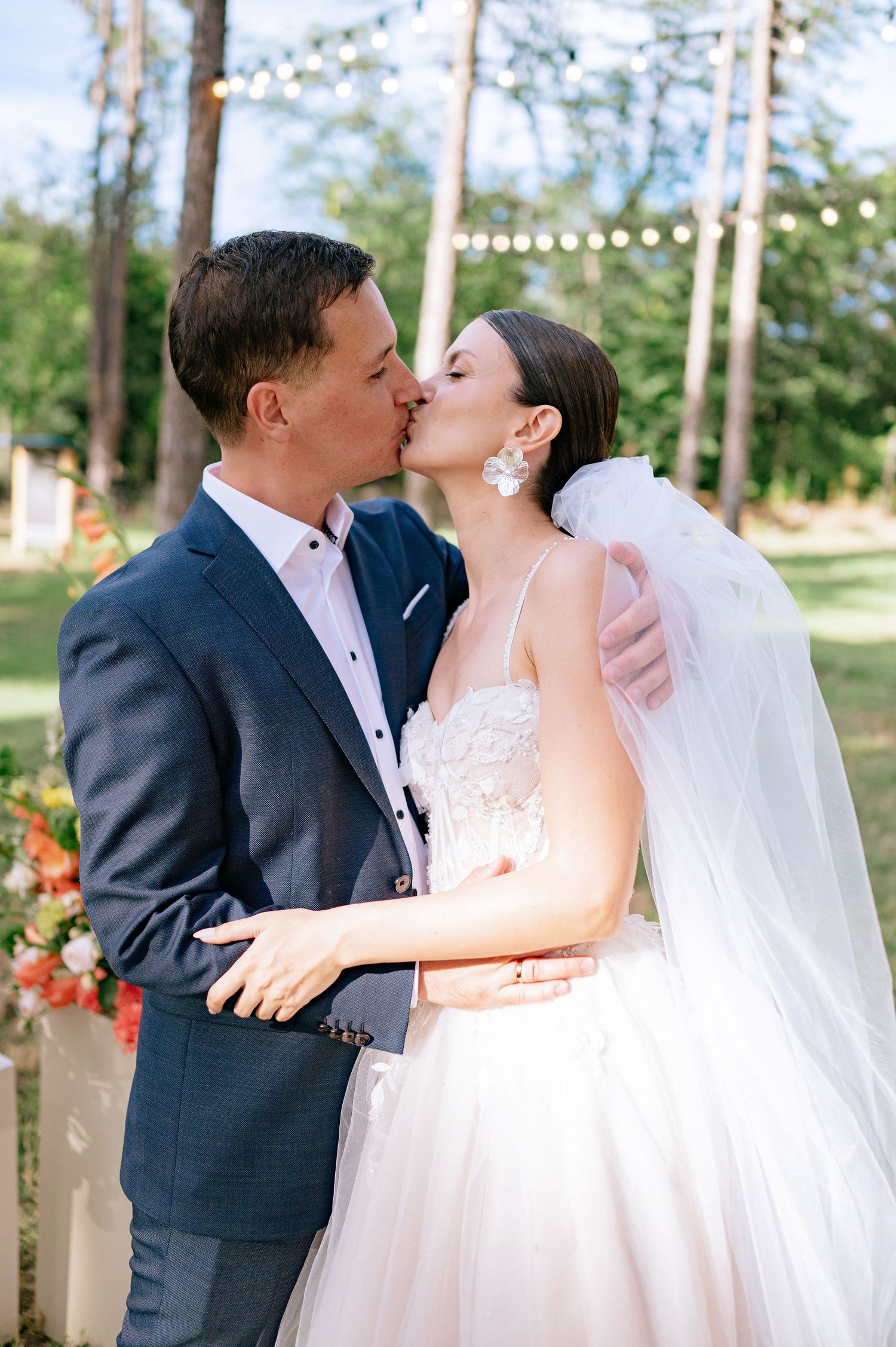 Newlyweds share a romantic kiss in an outdoor garden wedding setting with string lights and floral decorations.