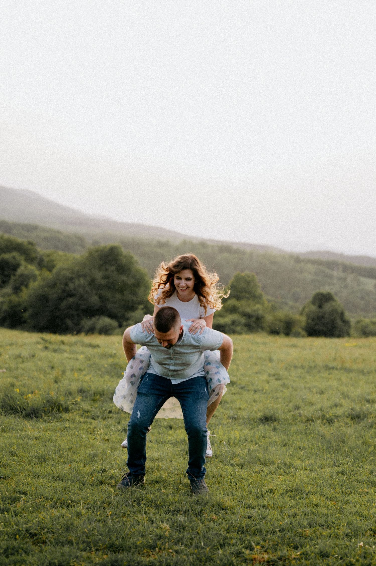 A couple plays and laughs together in a sunlit field with rolling hills in the background.