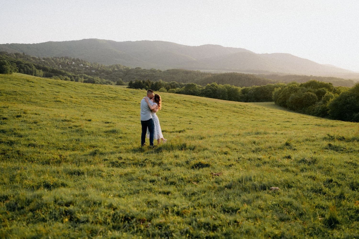 Two people embrace while standing in a sunlit mountain meadow surrounded by rolling hills.