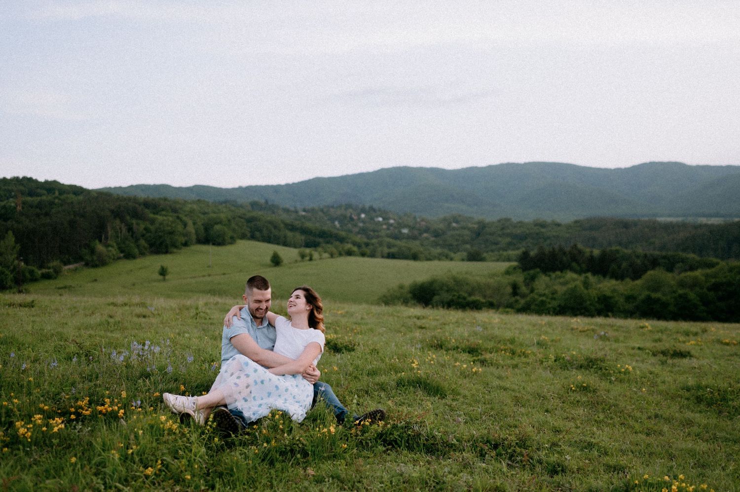 Two people share an intimate moment while sitting in a grassy mountain field with distant hills.