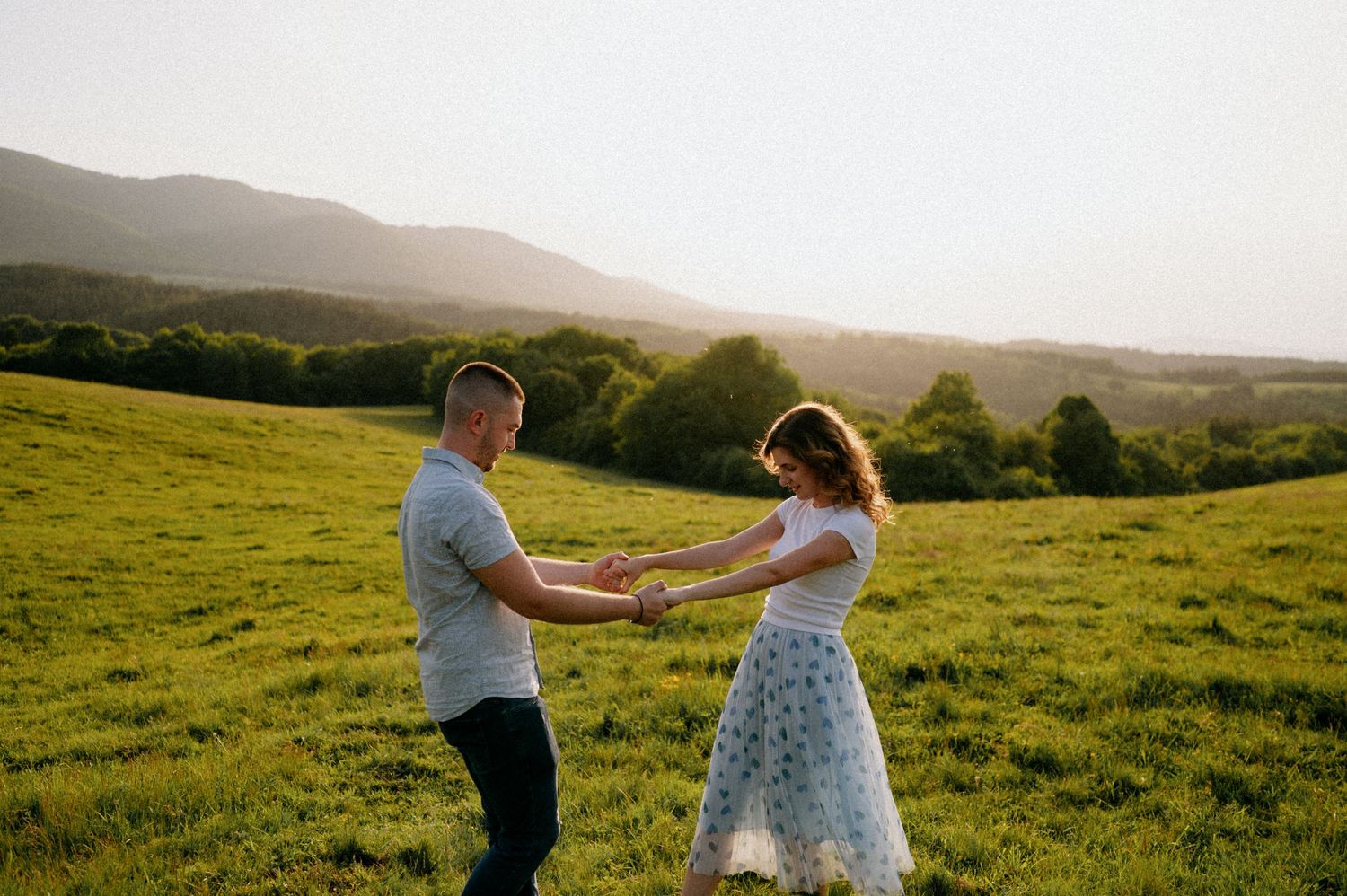 Couple enjoying a romantic dance in a sunlit meadow with mountains in the distance.