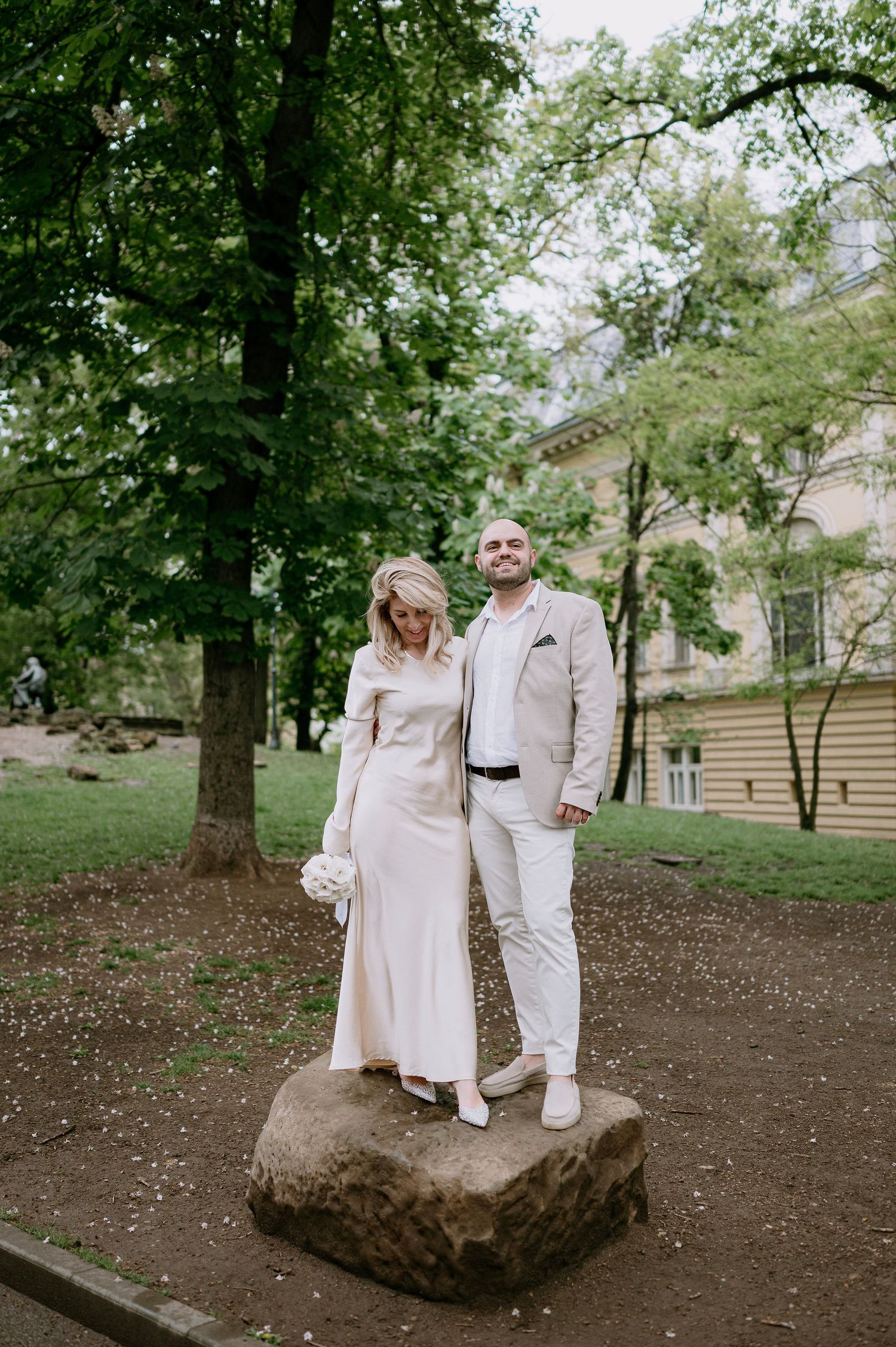 A couple in cream-colored clothing shares a moment together on a large rock in a park.