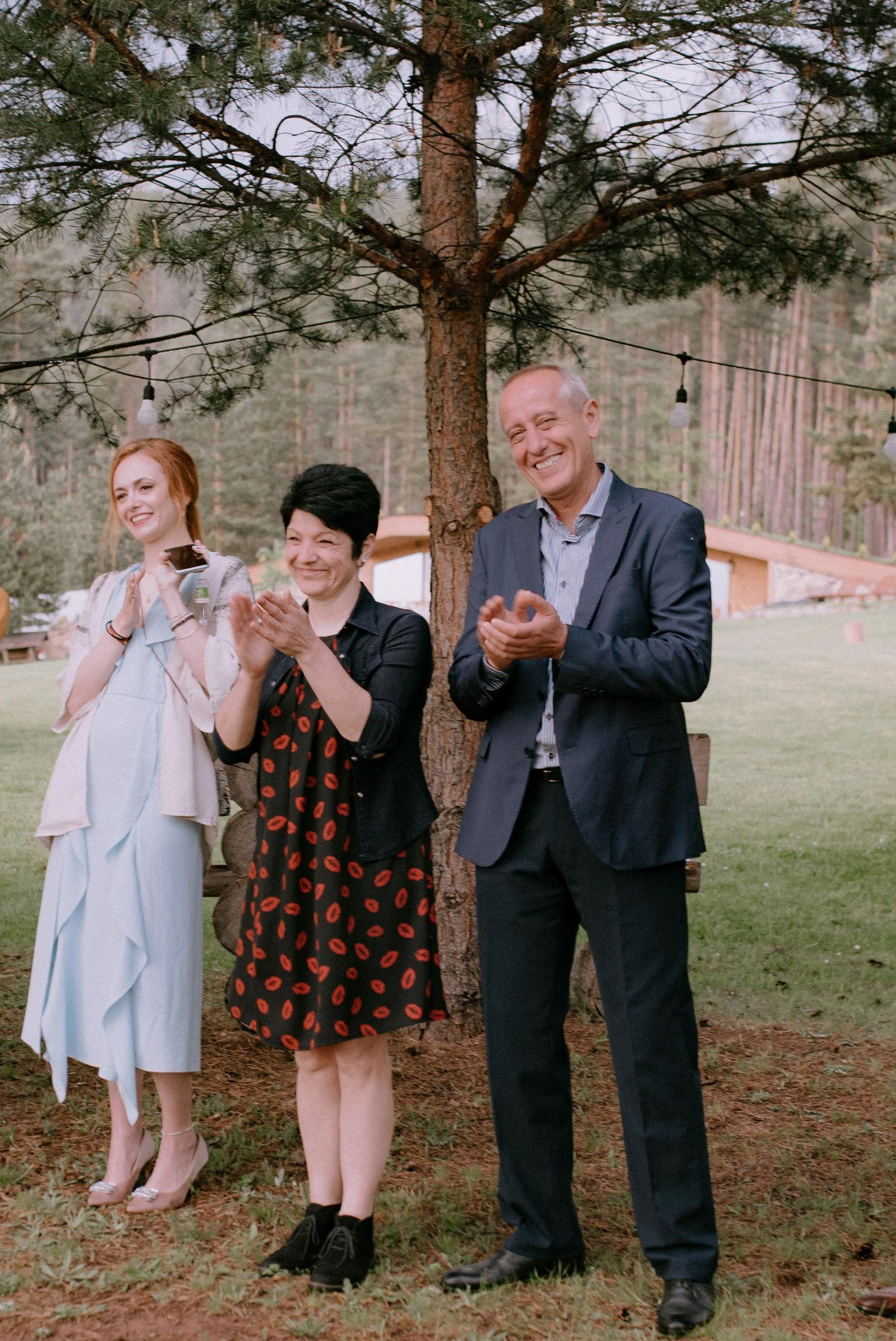 Group poses playfully under string lights and pine trees at outdoor wedding.