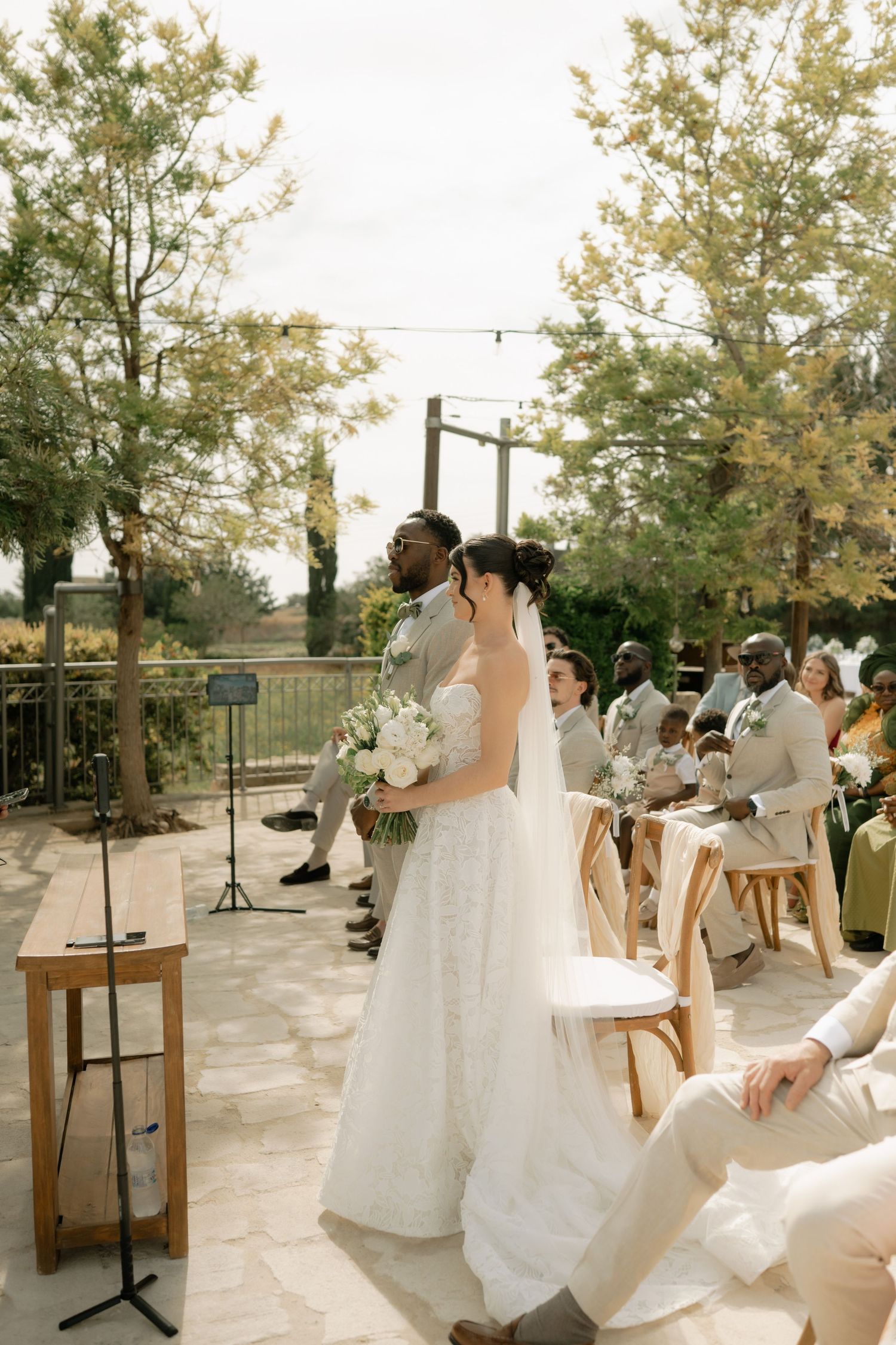 Bride and groom stand at outdoor wedding ceremony aisle, surrounded by guests seated in wooden chairs amid lush greenery.