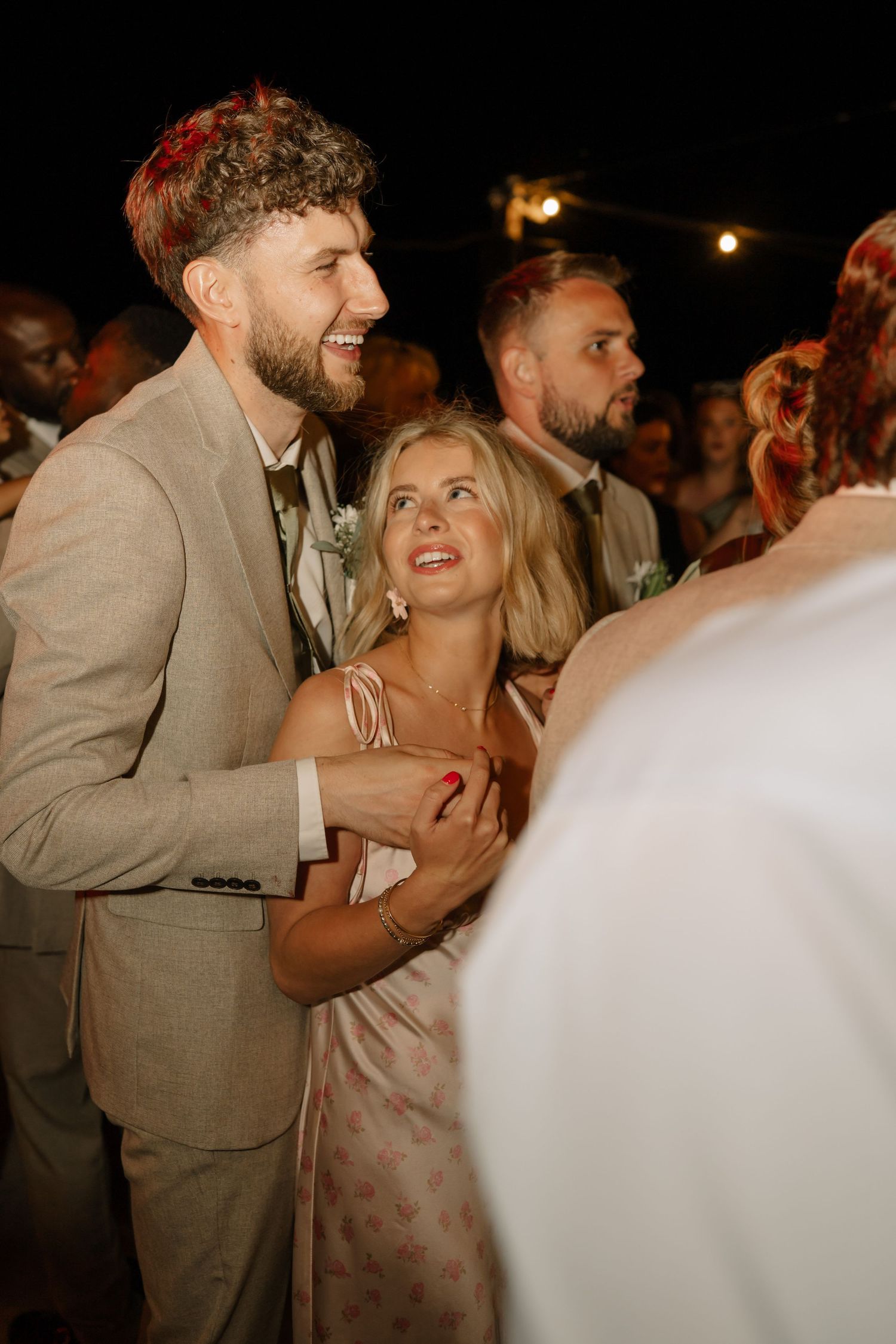 A couple in formal attire laughs joyfully at an outdoor evening wedding reception surrounded by guests.