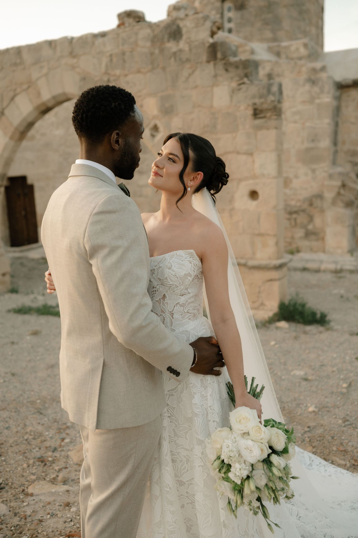 Interracial couple in wedding attire embrace before ancient stone ruins, bride holding white bouquet.
