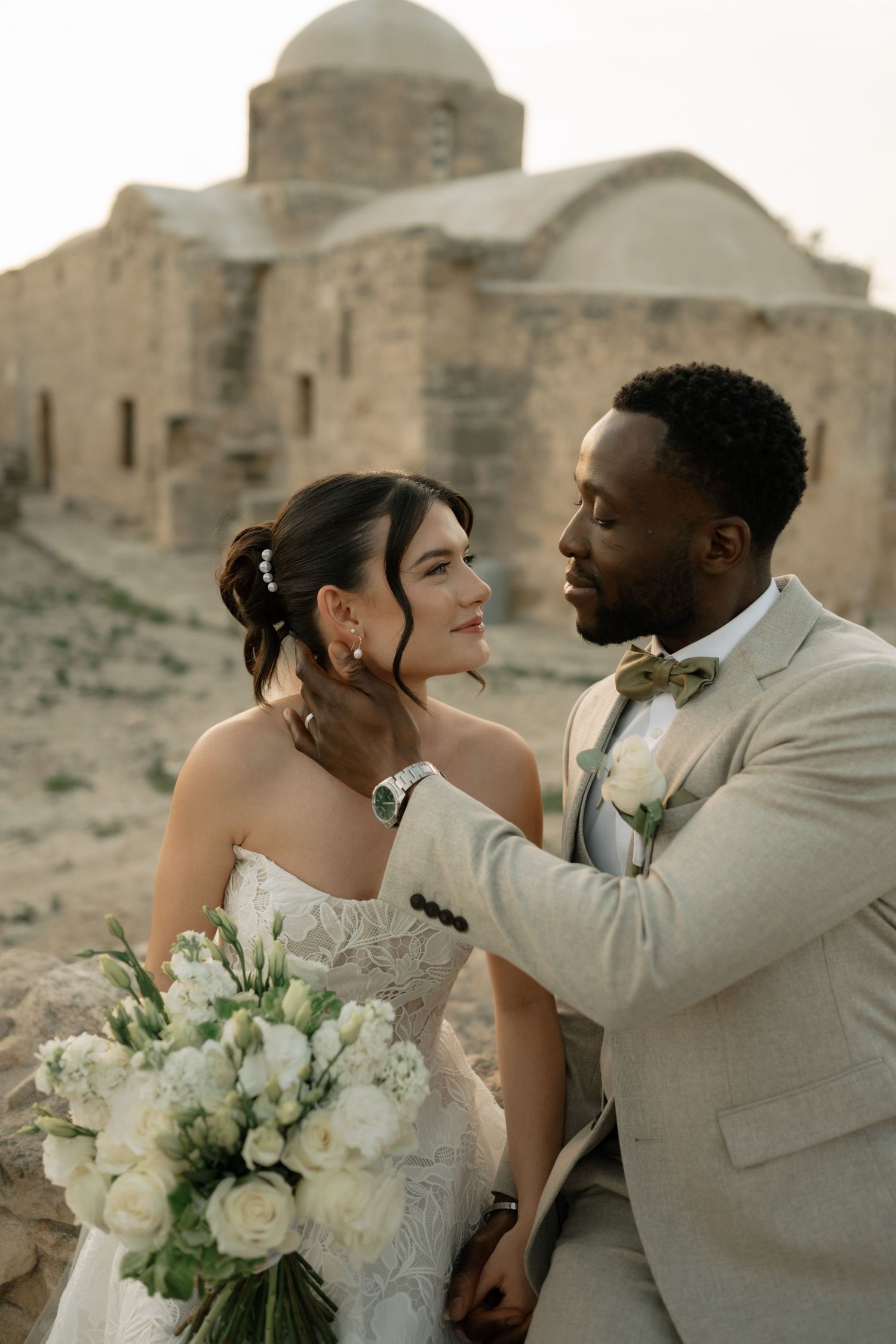 Interracial couple in wedding attire pose romantically before ancient stone church ruins in arid landscape.