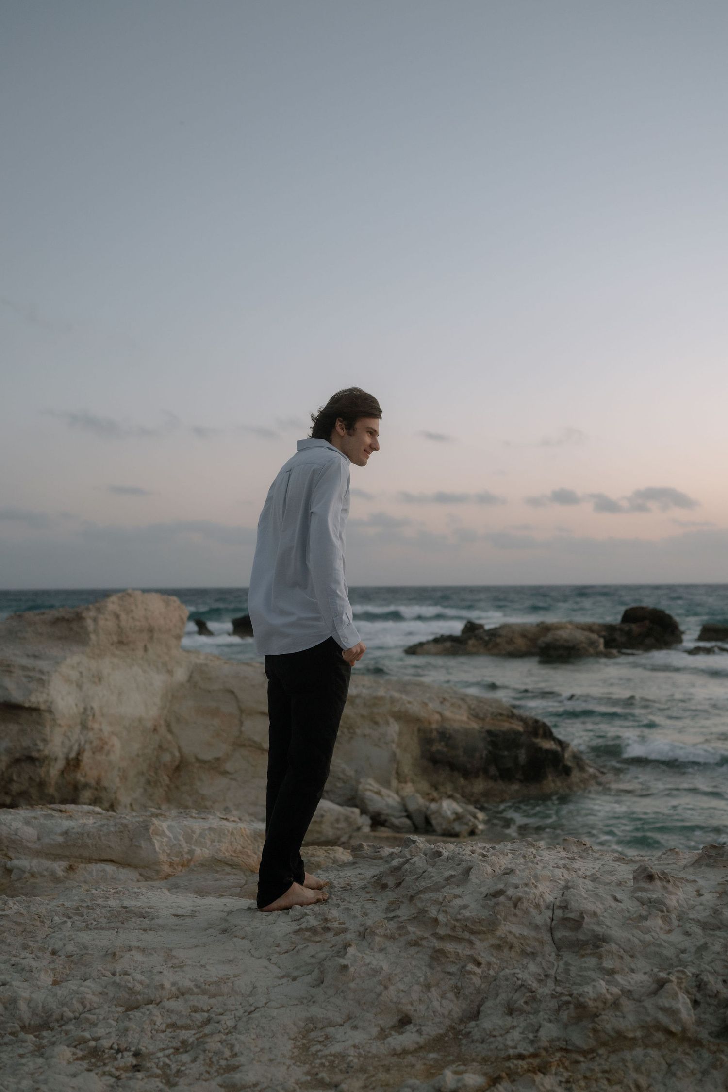 Young man in white shirt and black pants standing barefoot on rocky shoreline at dusk with ocean waves behind him.