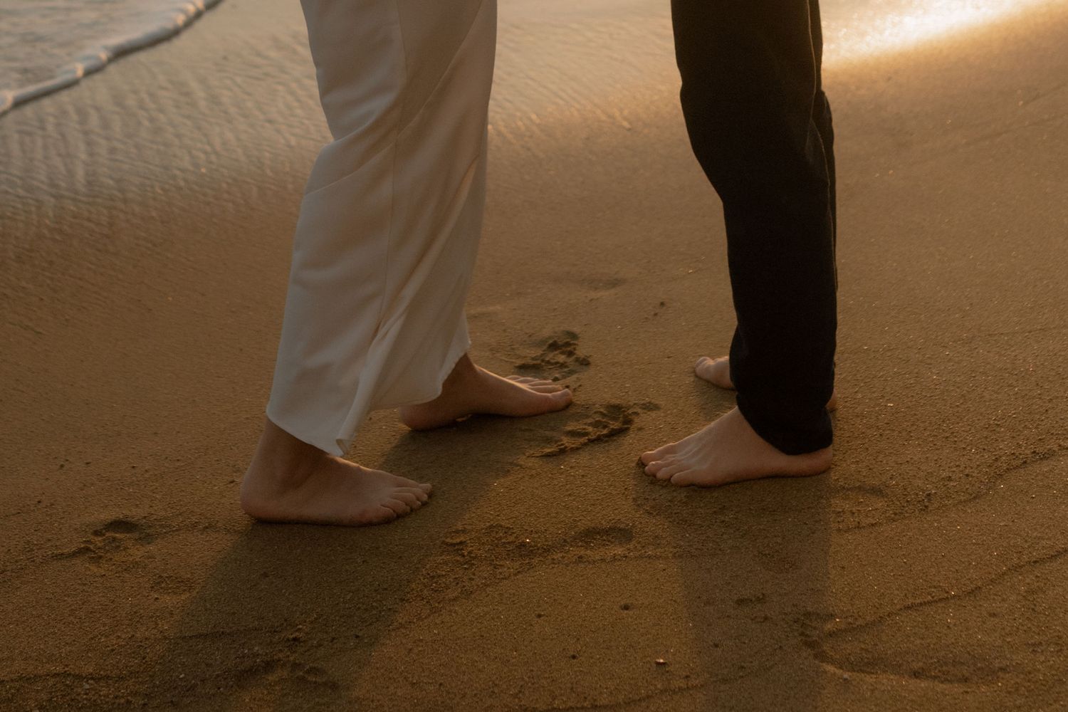 Two barefoot people standing on a sandy beach at sunset, leaving footprints in the wet sand.
