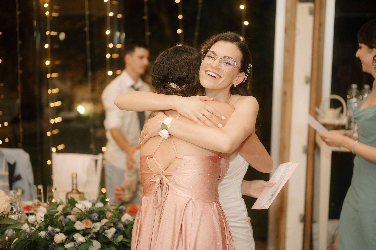Wedding guests in peach-colored dresses embrace during an evening reception with twinkling lights in the background.