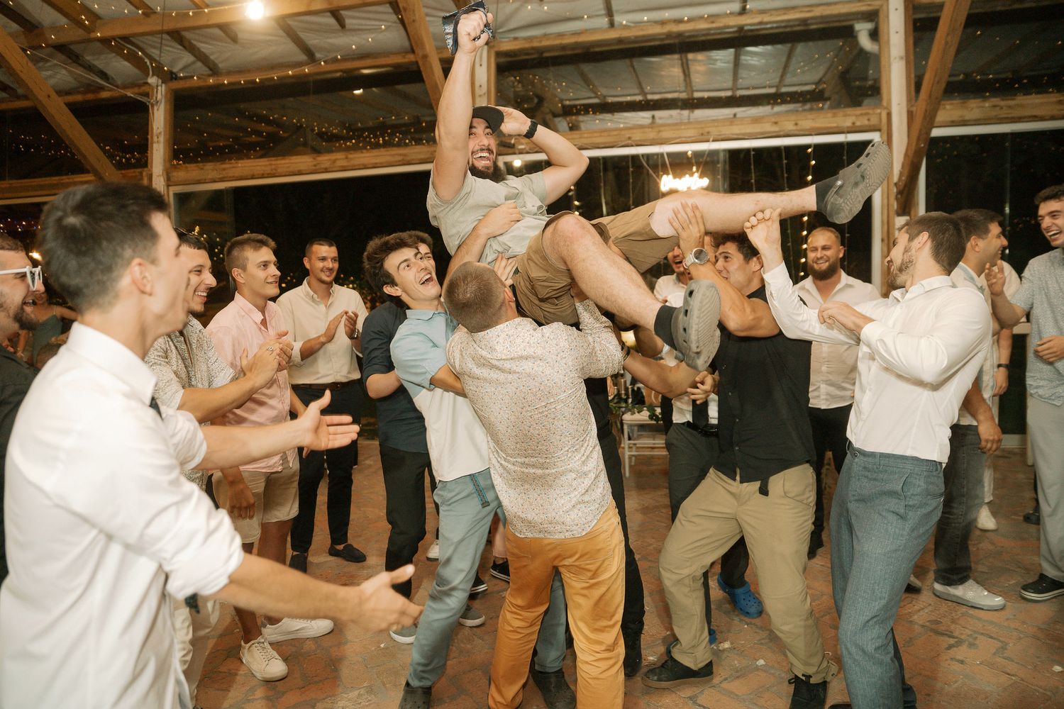 A large group of wedding guests dance together in a line formation on a wooden floor at an indoor reception venue.