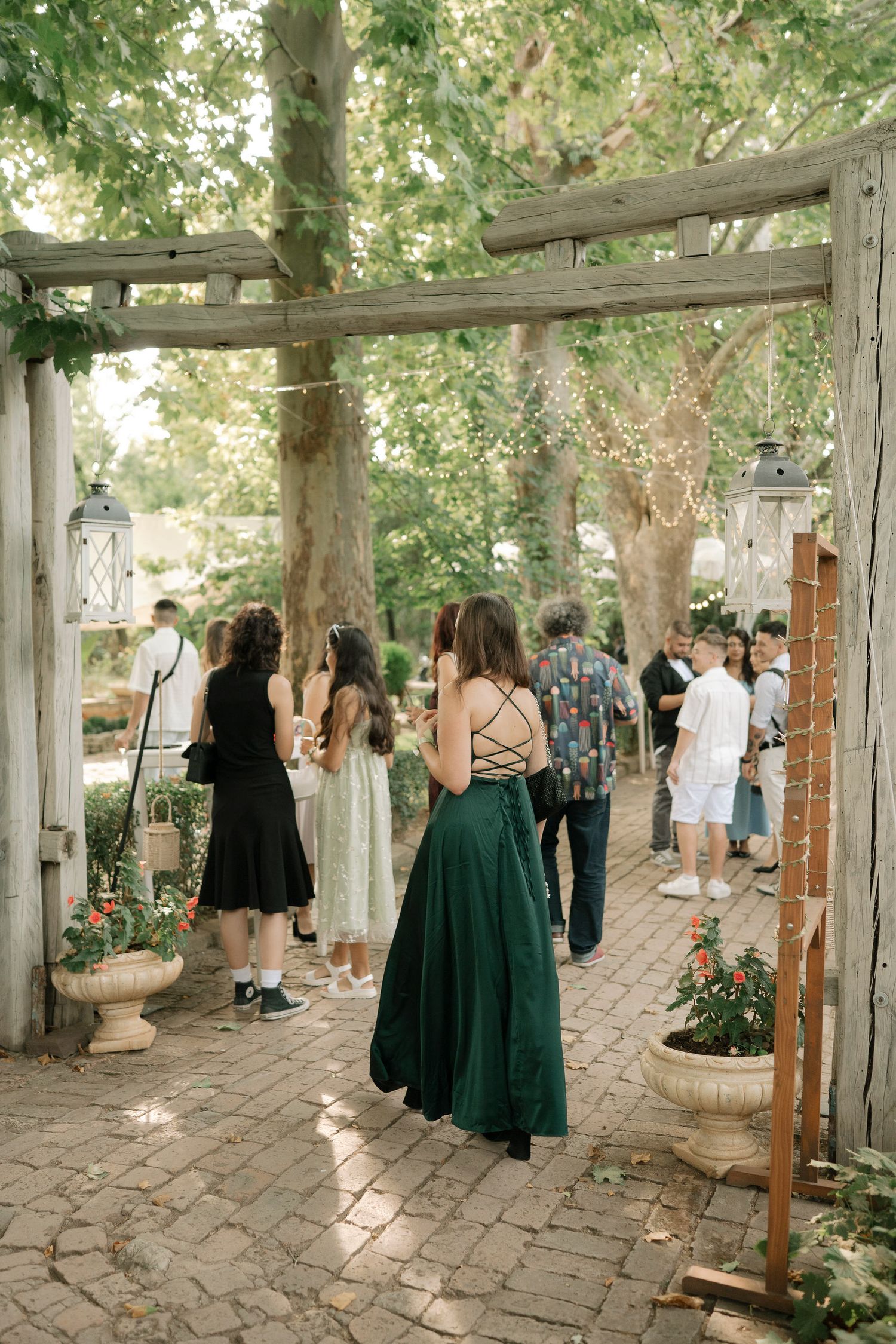 Guests gather in a rustic outdoor venue with wooden pergolas and stone pathways during a wedding ceremony.