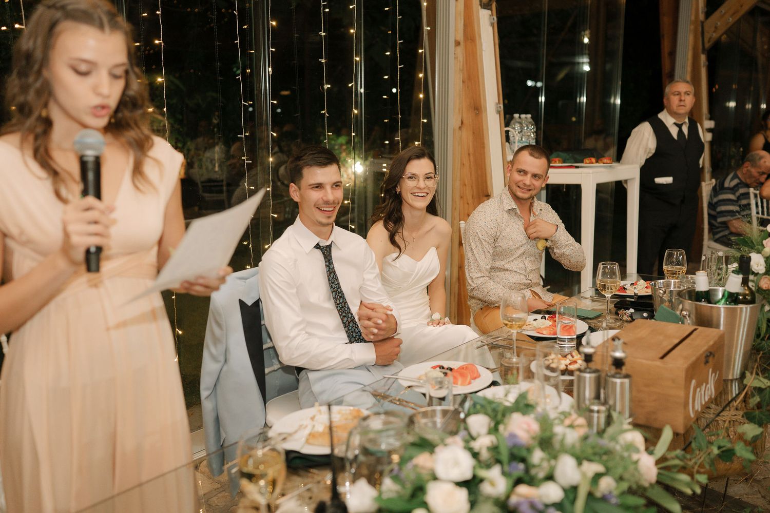 Wedding reception guests seated at long tables decorated with greenery and candles in an elegant glass-enclosed venue.
