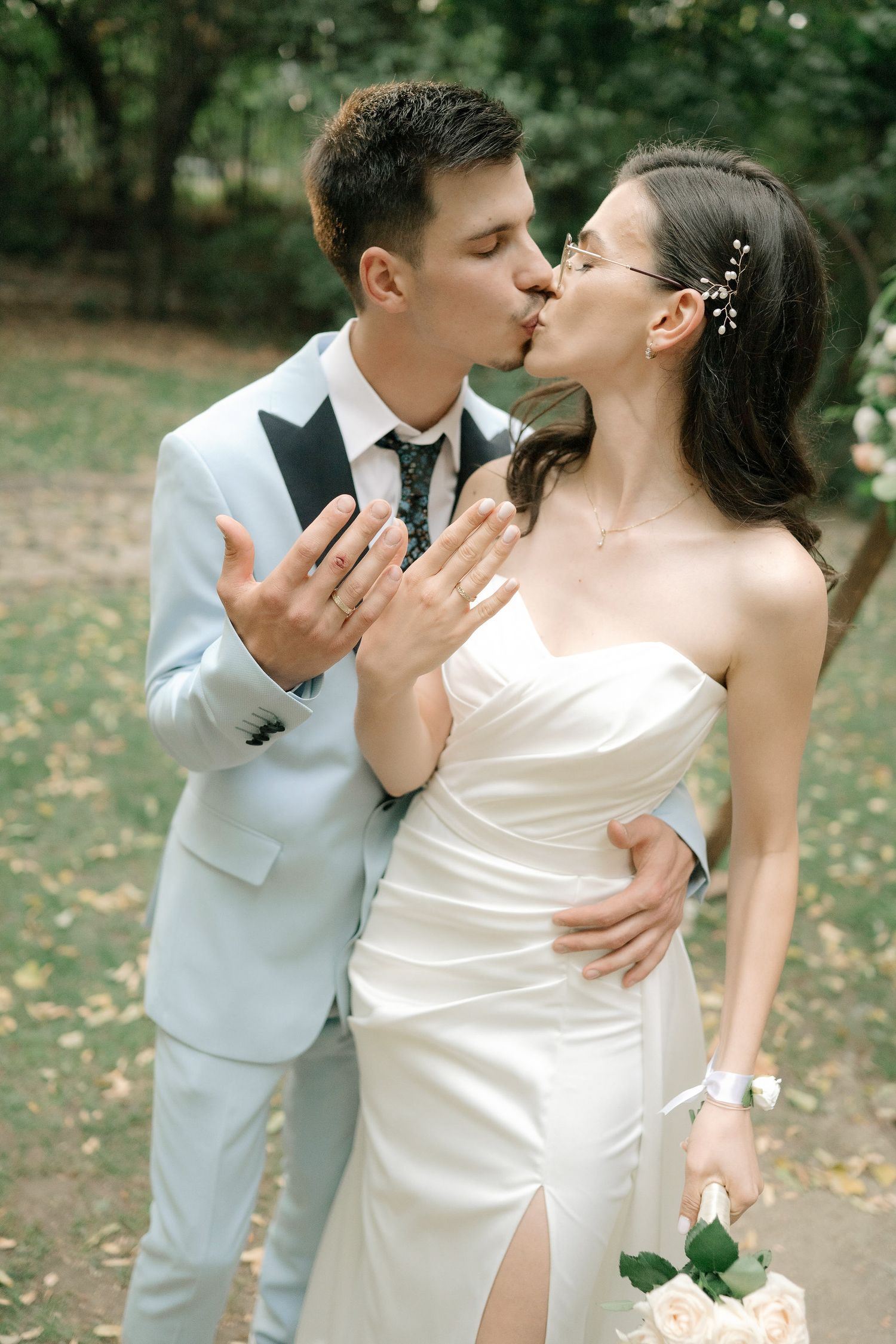 A romantic wedding couple embraces and shares a tender kiss in an outdoor garden setting with fallen autumn leaves.