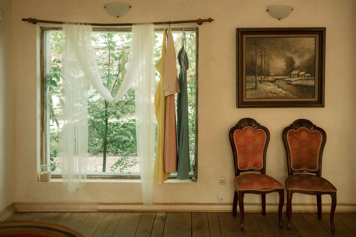 Vintage chairs and framed artwork sit beside a window with sheer curtains in a rustic room with wooden floors.