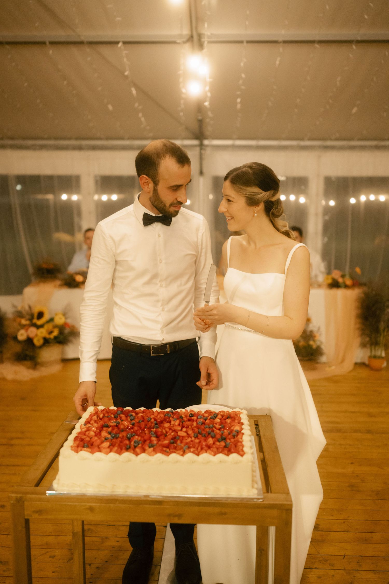 A couple in wedding attire arranges a long dessert display table with strawberries and other treats at their reception.