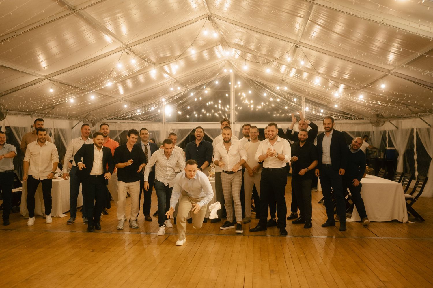 Wedding guests posing together on a wooden dance floor in an elegant tent with string lights overhead.