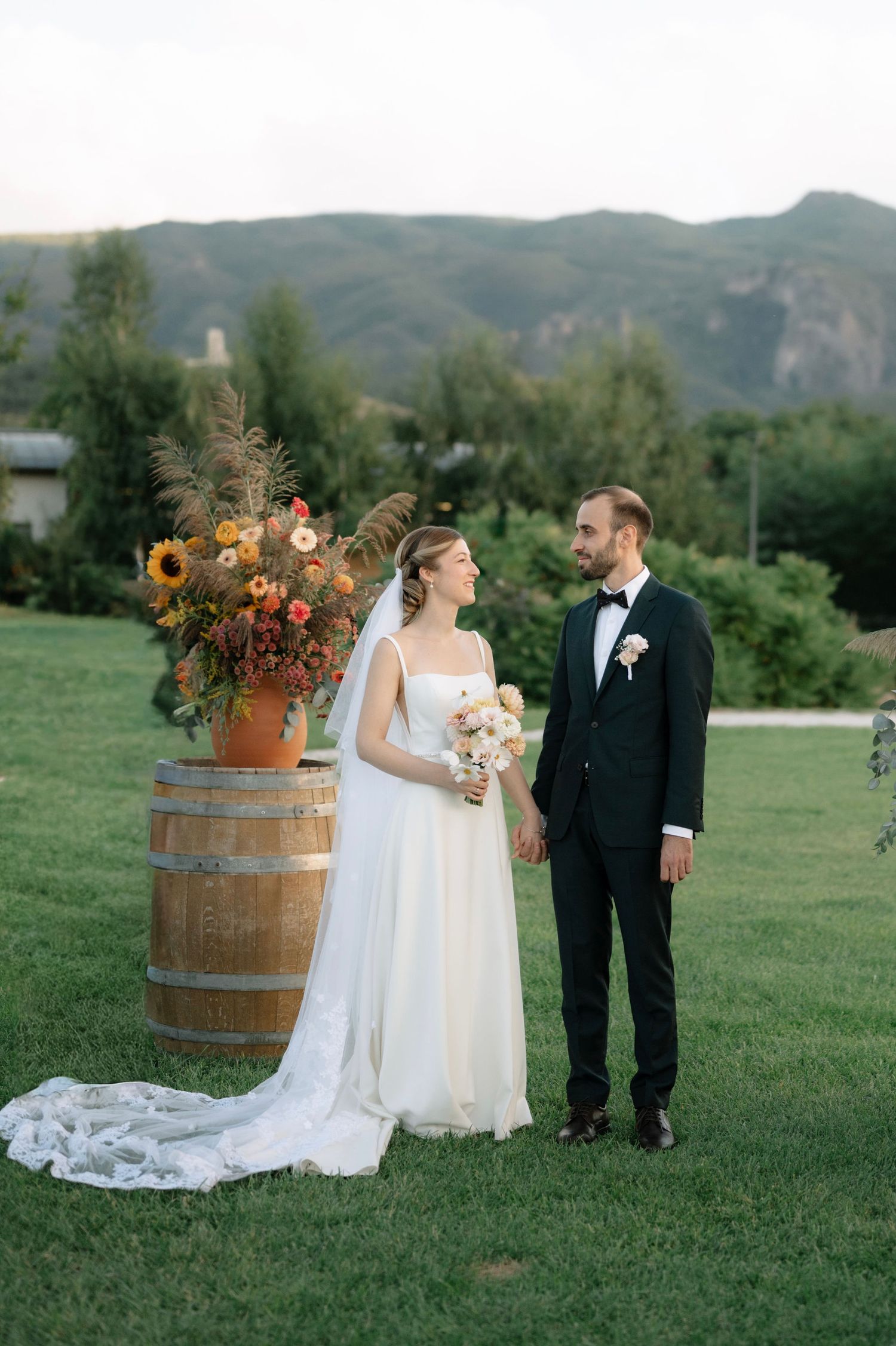 Elegant outdoor wedding couple poses in front of rustic wine barrels on a scenic grassy hillside.