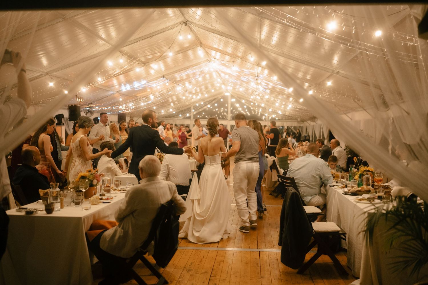 Wedding reception guests seated at tables under twinkling string lights in an elegant white tent.