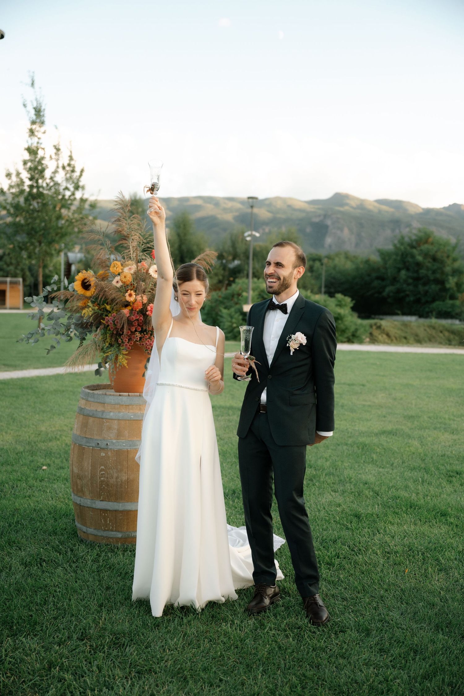 A rustic outdoor wedding scene with a wine barrel and sunflower decor against rolling hills in the background.