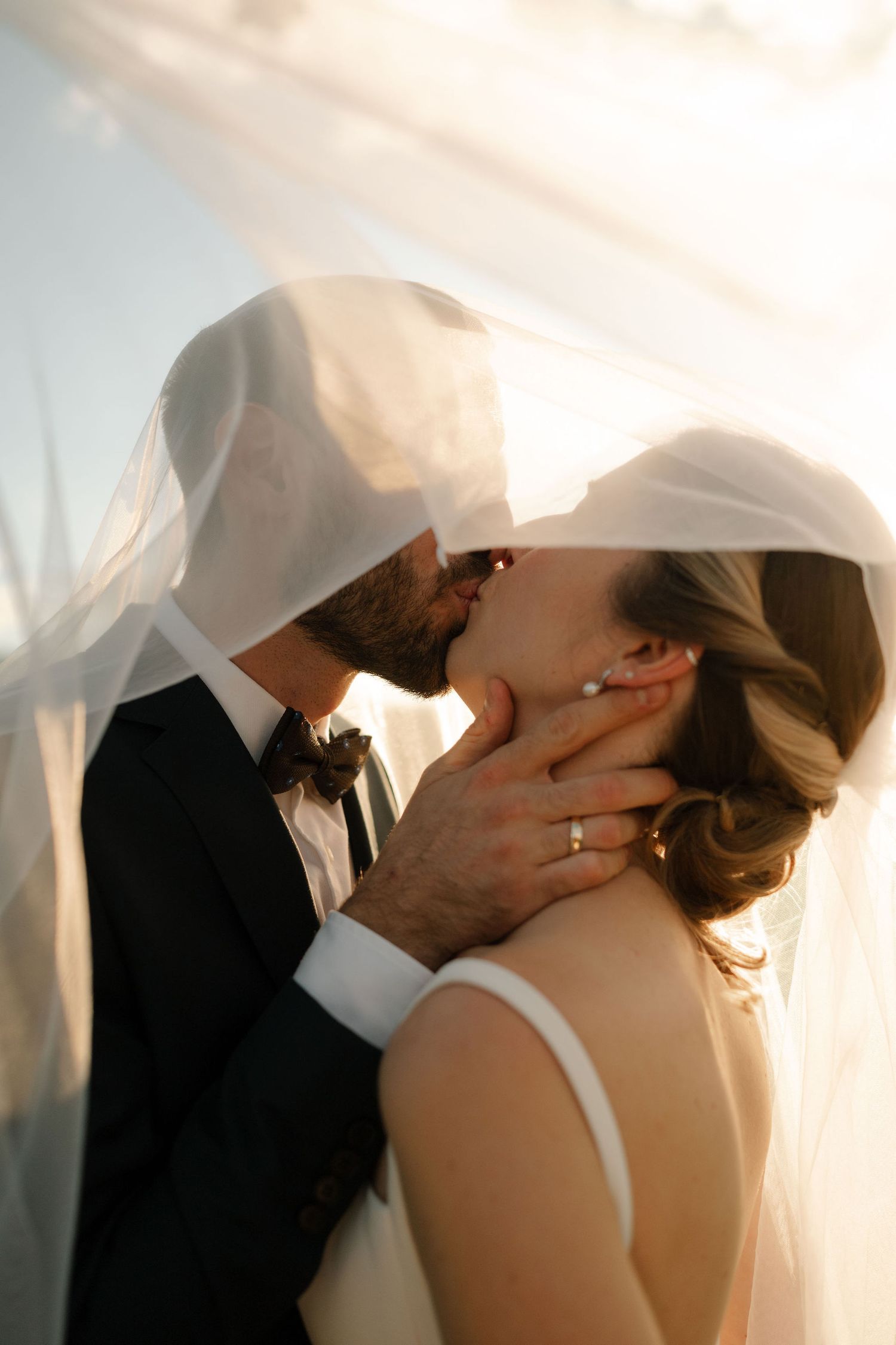 A romantic wedding couple shares an intimate kiss under a flowing white veil during their outdoor ceremony.