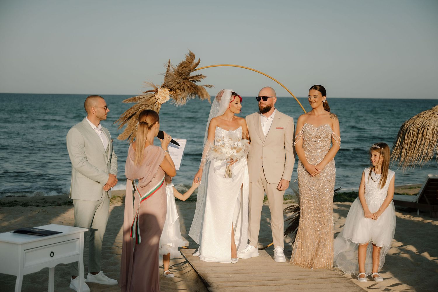 A beachside wedding ceremony takes place on a wooden deck by the ocean as wind blows through decorative palm fronds.