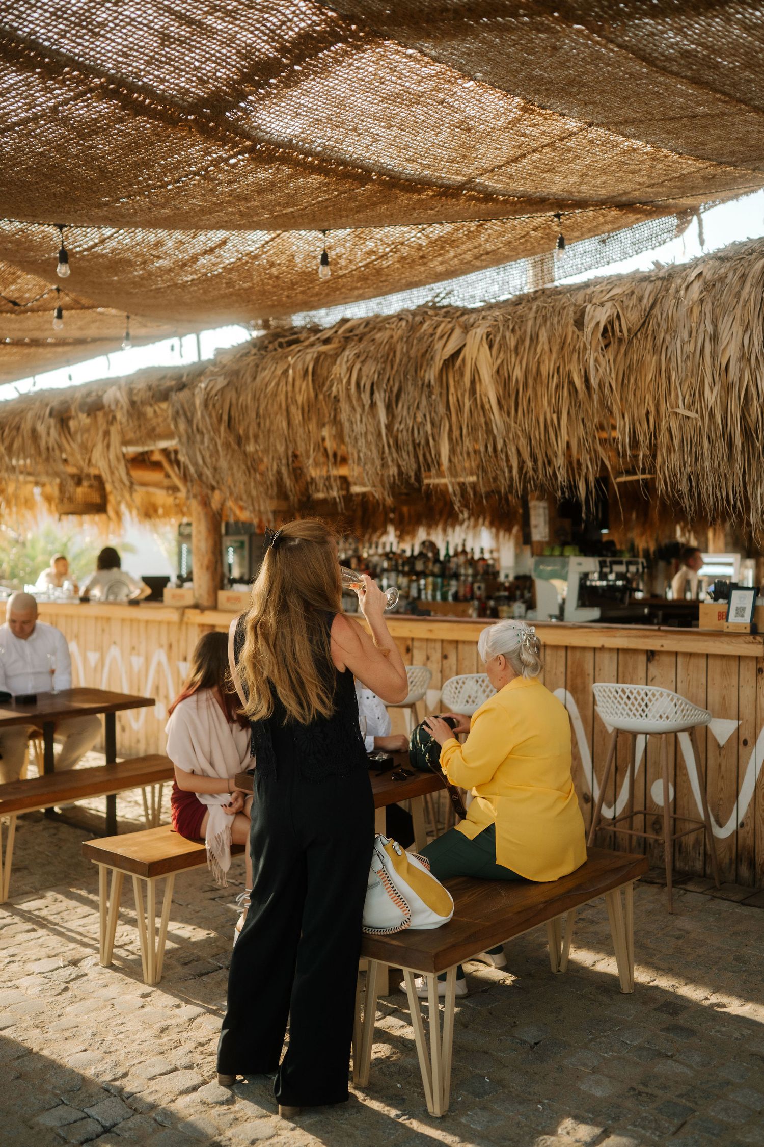Patrons enjoy drinks at a rustic beach bar with thatched palm roof and wooden furniture in warm sunlight.