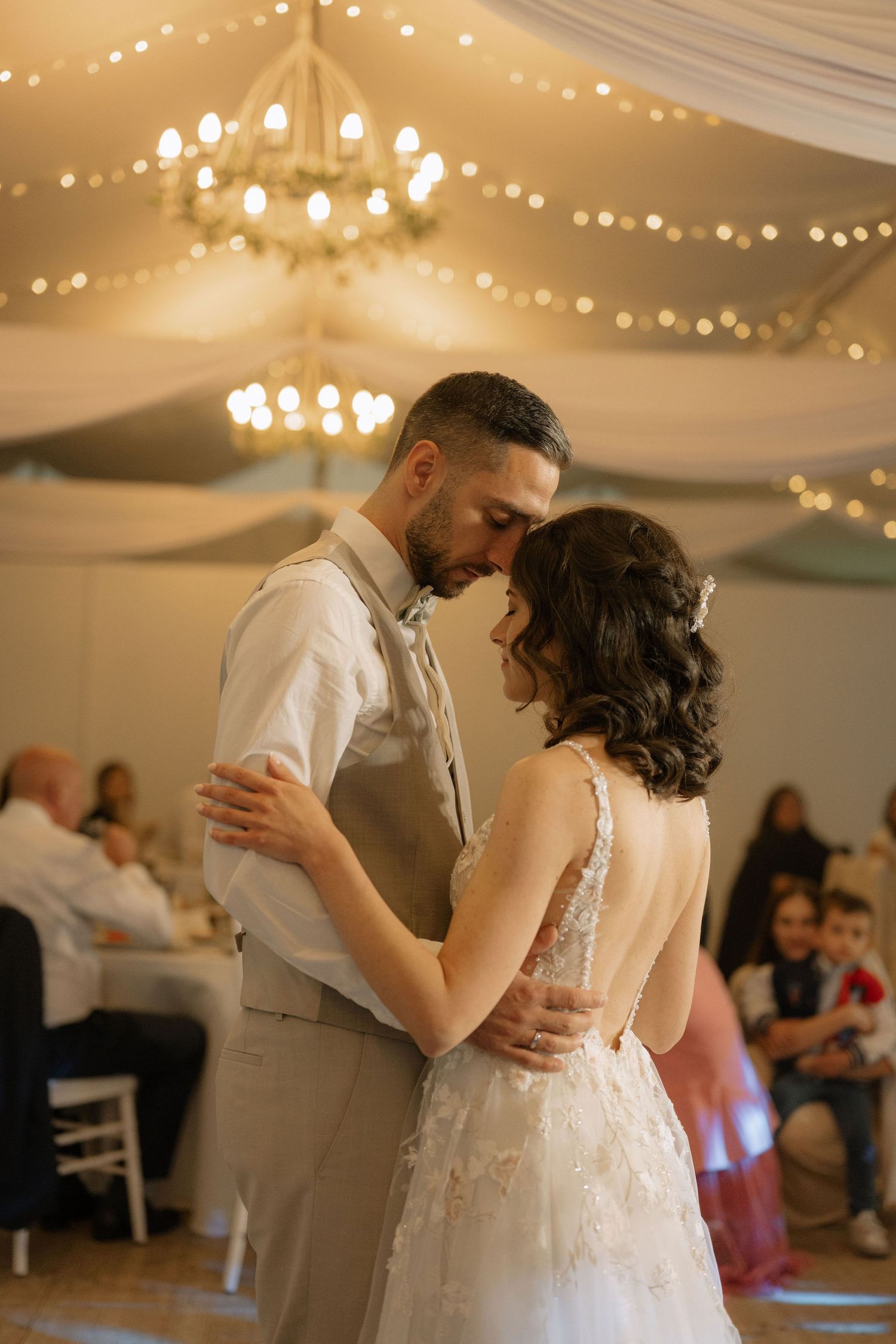 Bride and groom share their first dance under a chandelier with warm fairy lights in an elegant wedding reception tent.